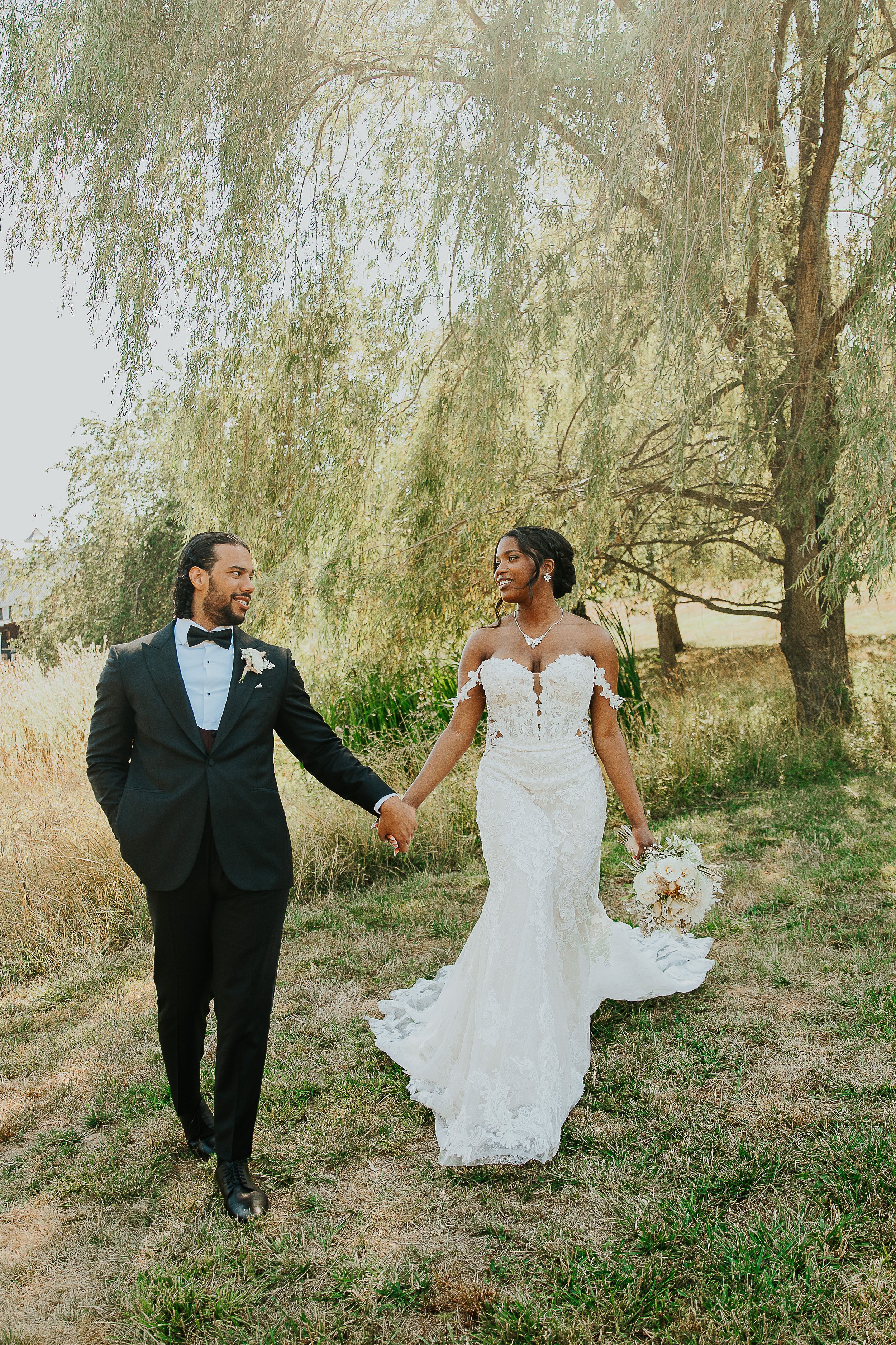 A bride and groom holding hands, walking outdoors on a grassy area with trees in the background. The groom is wearing a black tuxedo with a bow tie, and the bride is in a white lace wedding dress holding a bouquet of flowers.
