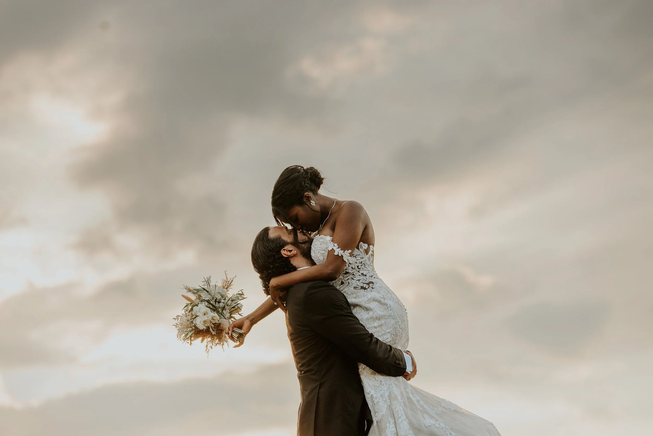 A newlywed couple dancing outdoors under a cloudy sky. The groom lifts the bride, who holds a bouquet, and they gaze at each other.