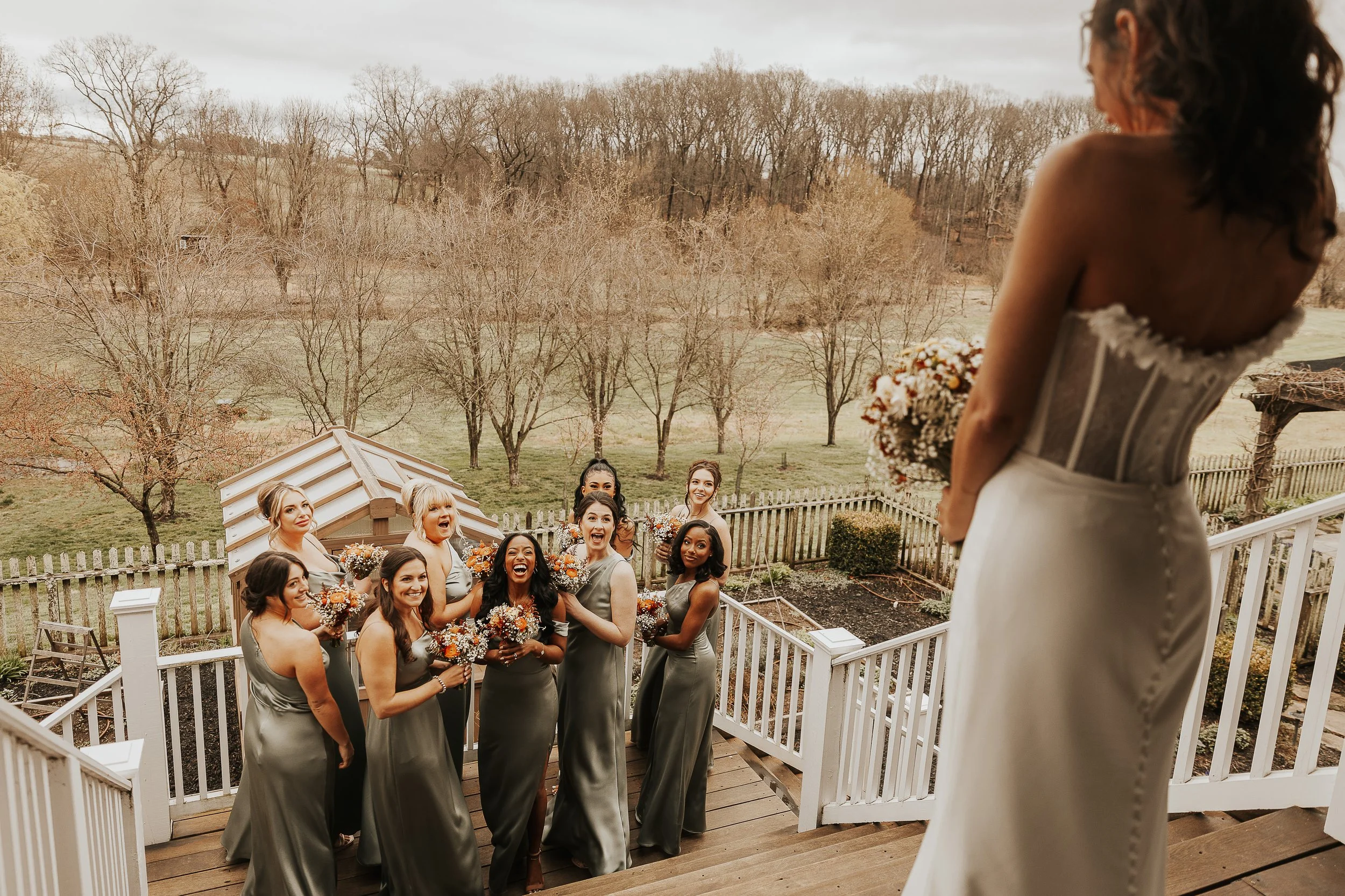 A bride facing her bridesmaids on a backyard porch with leafless trees in the background.