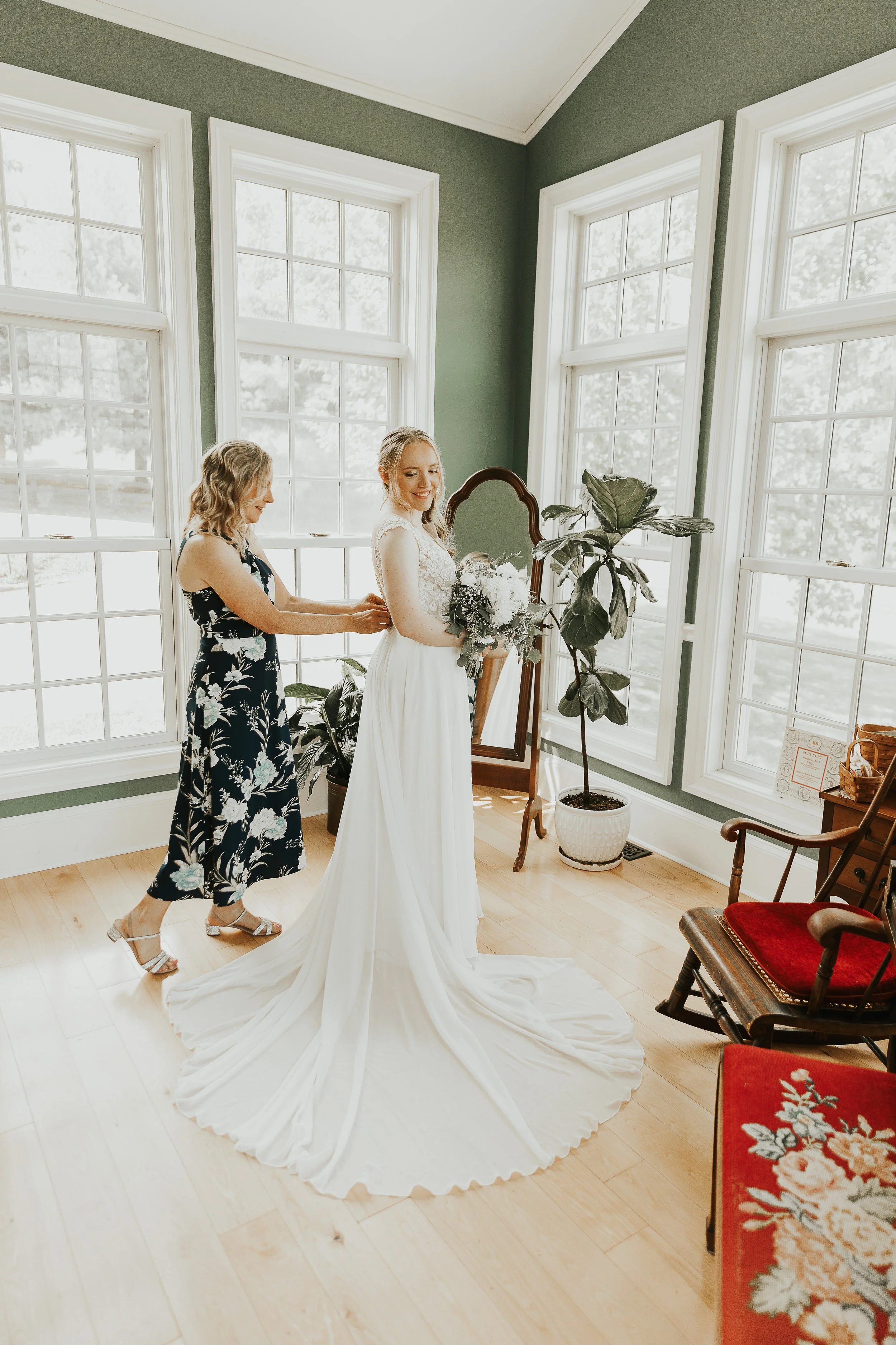 A bride in a white wedding dress is smiling with her eyes closed while a woman, possibly her mother, helps her with her dress or accessories in a bright room with large windows, green walls, and wooden floors. There are potted plants, a mirror, and r