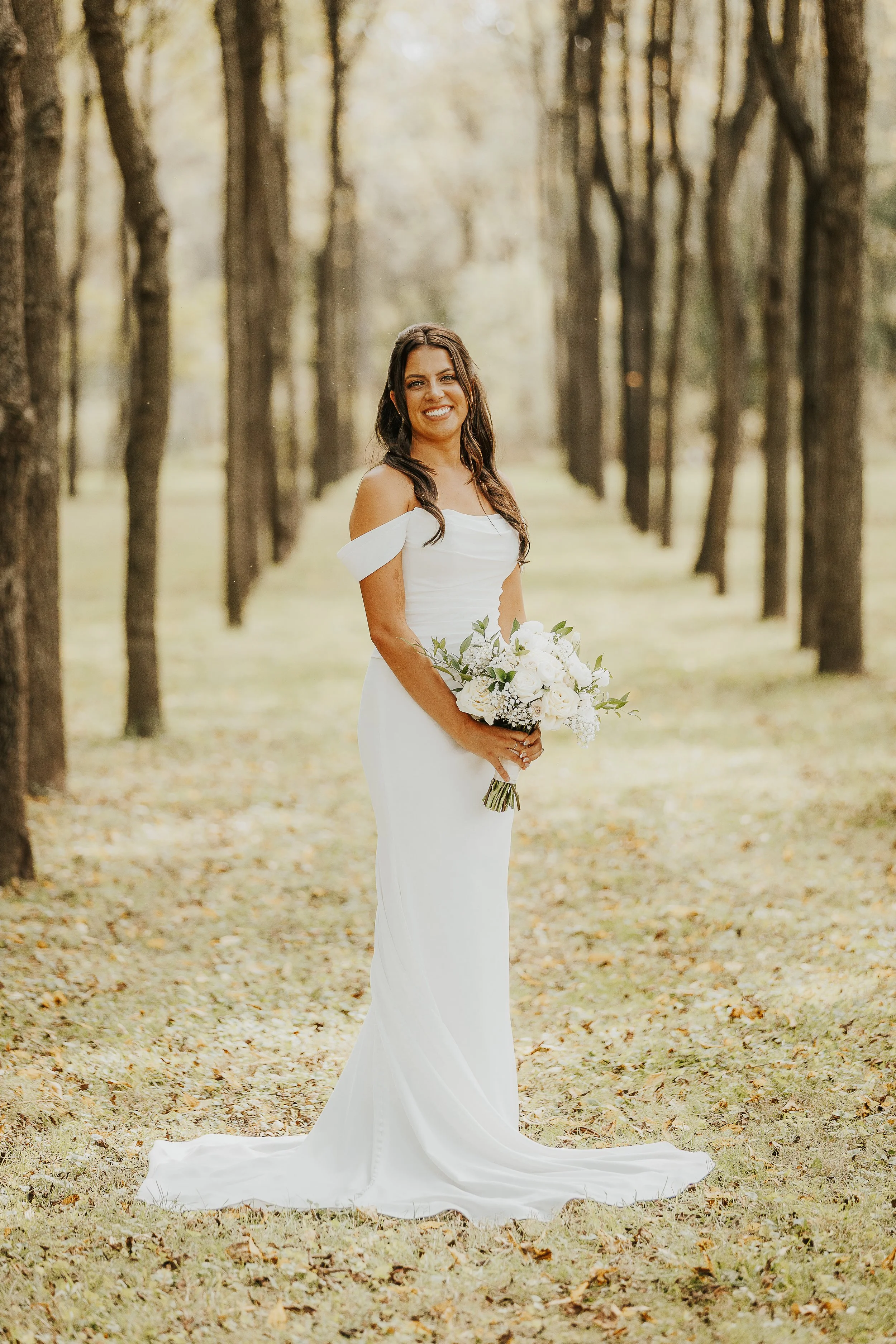A woman in a white off-shoulder wedding dress standing in a tree-lined outdoor setting with fallen leaves, holding a bouquet of white flowers, and smiling at the camera.