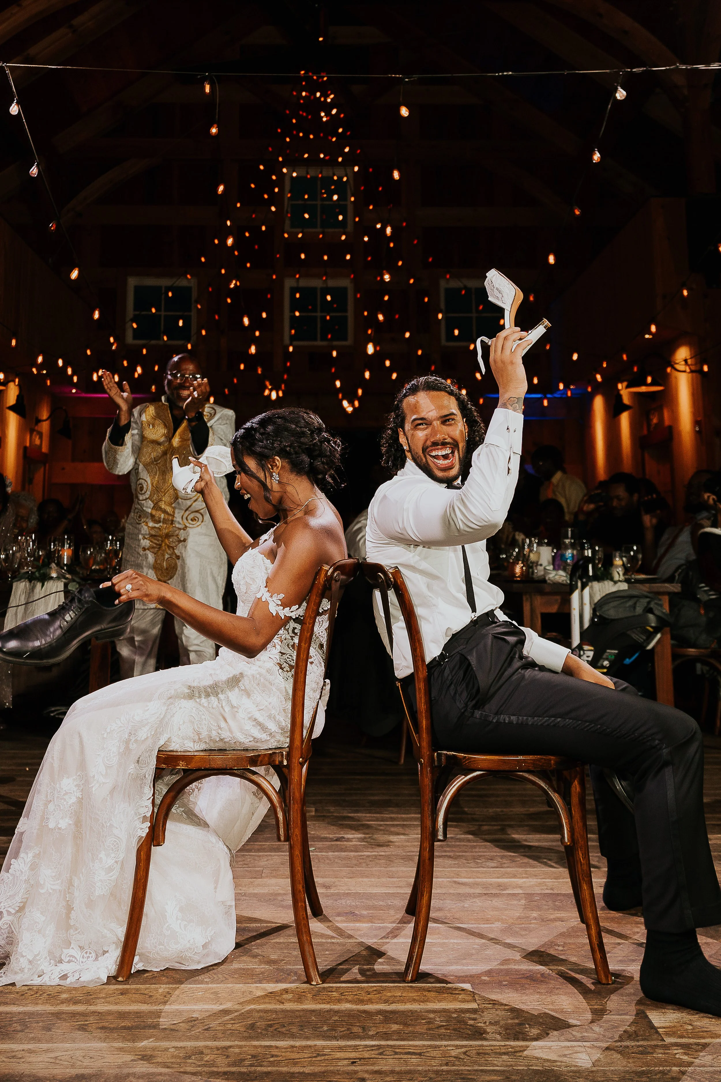 A wedding reception with a bride in a white lace wedding gown and a groom in a black suit and white shirt, sitting back to back on wooden chairs, smiling and celebrating. The bride is holding a shoe, and the groom is holding a shoe and a gift. A gues