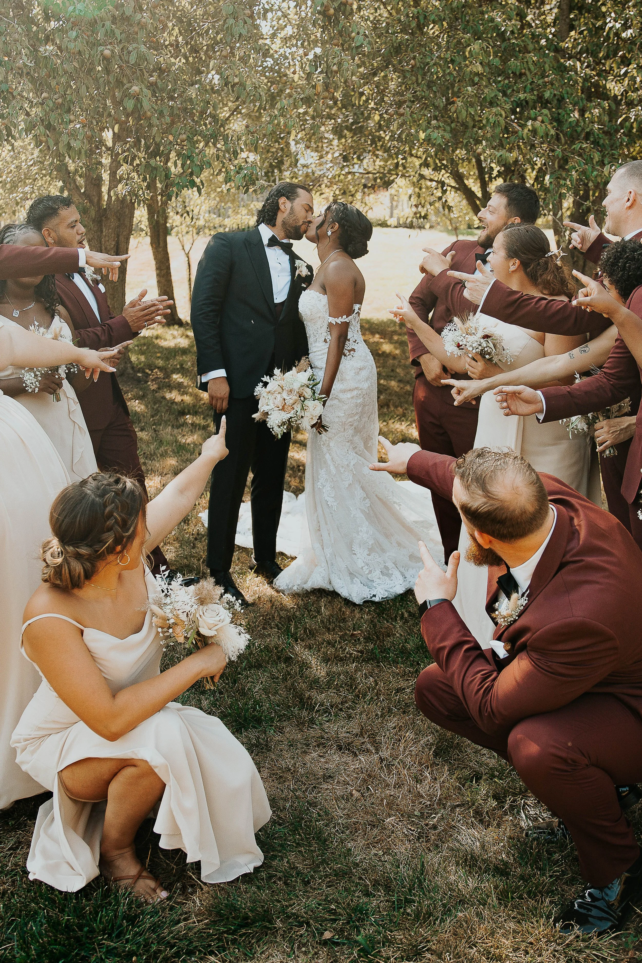 A group of wedding guests dressed in formal attire surrounds a bride and groom outdoors. The bride, in a white lace wedding dress, and the groom, in a black tuxedo, kiss in the center. The guests, in matching burgundy suits and beige dresses, point a