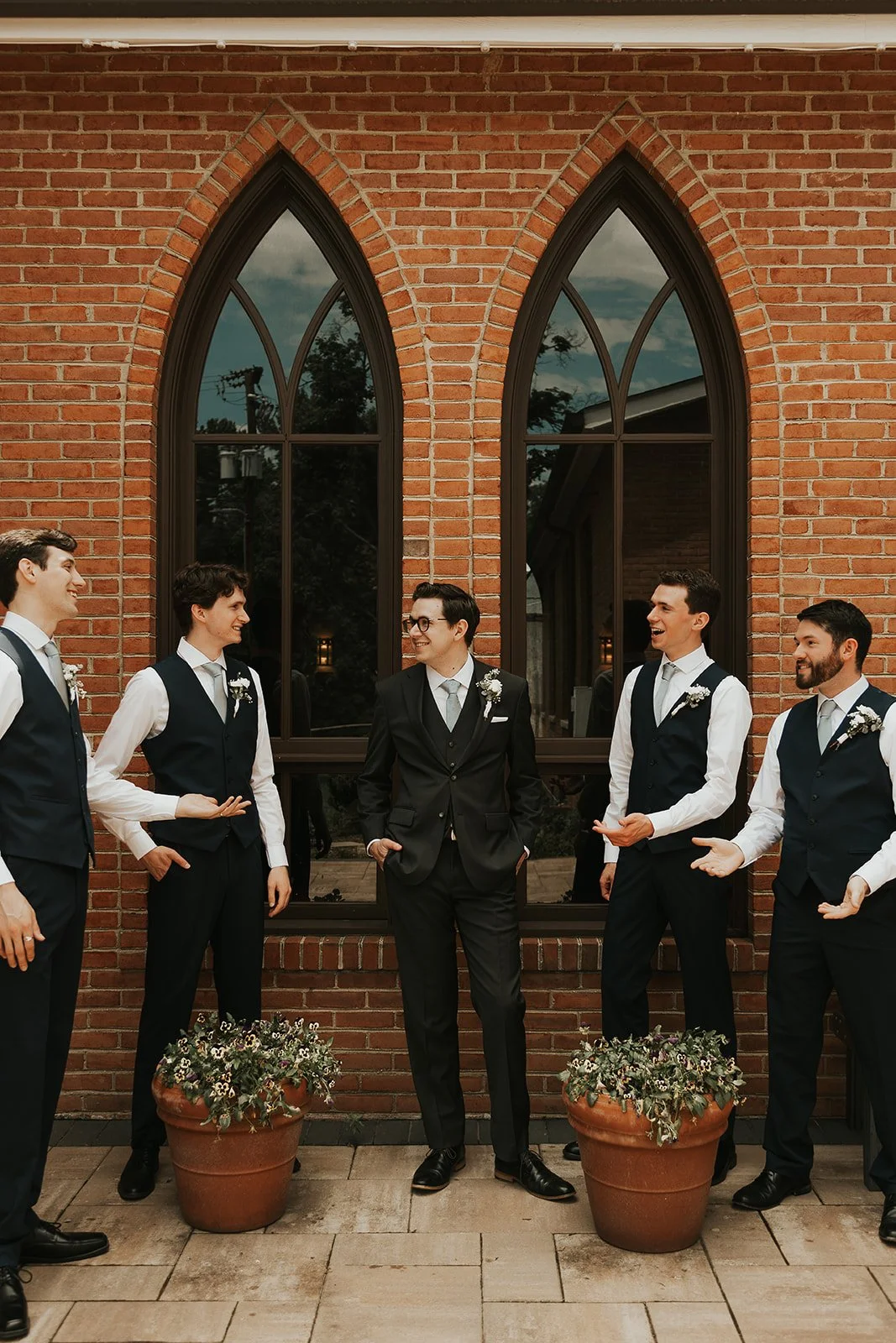 Group of five men dressed in formal suits and vests, standing and talking outside in front of a brick building with large Gothic-style arched windows, two potted plants with flowers in the foreground.