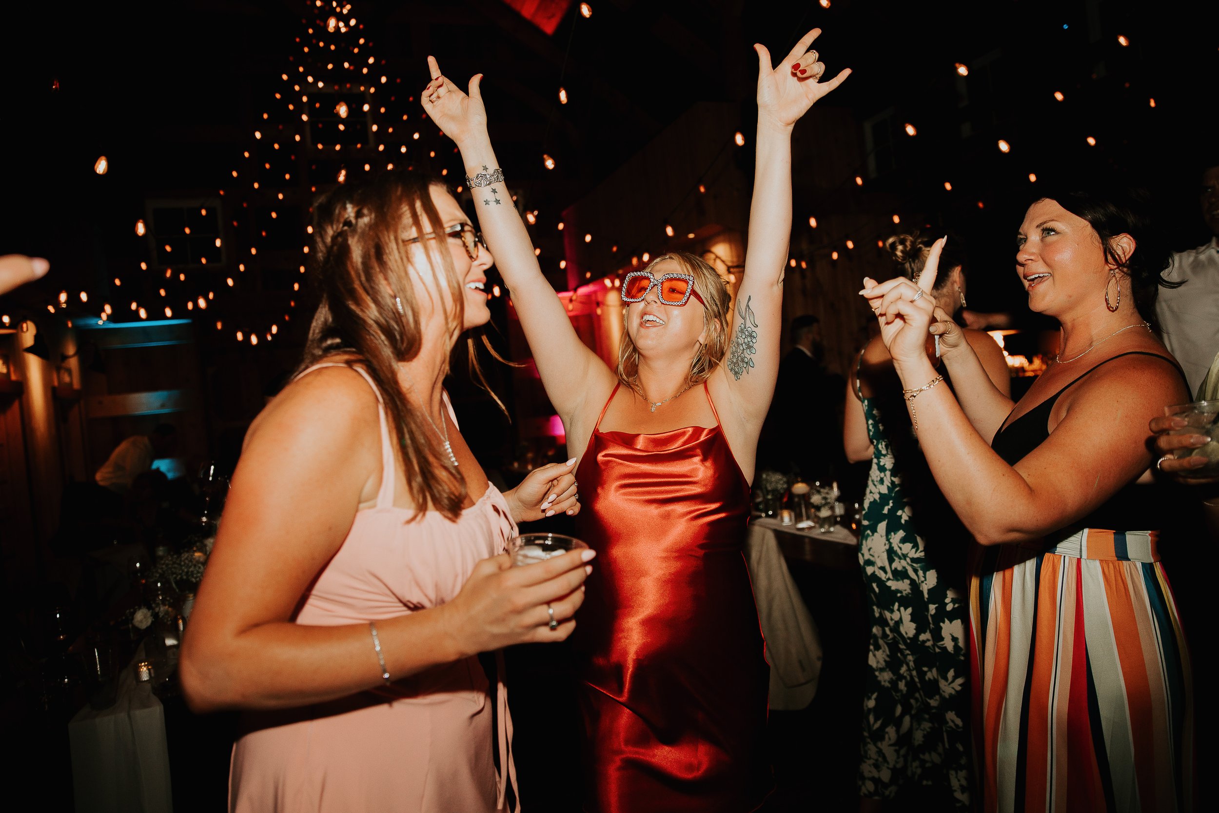 Group of women dancing and socializing at a party in a dimly lit venue with hanging string lights, some women wearing colorful and patterned outfits, one woman wearing red sunglasses, enjoying drinks and music.