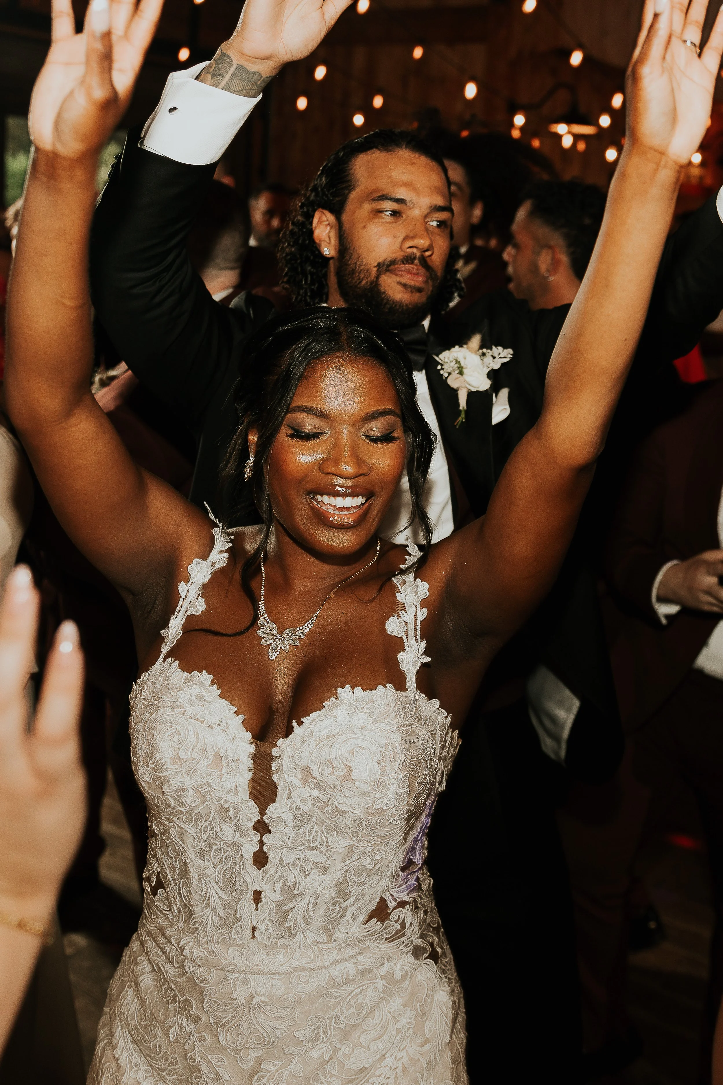 A bride in a white lace wedding dress and a necklace, celebrating with arms raised, smiling with eyes closed at a wedding reception.
