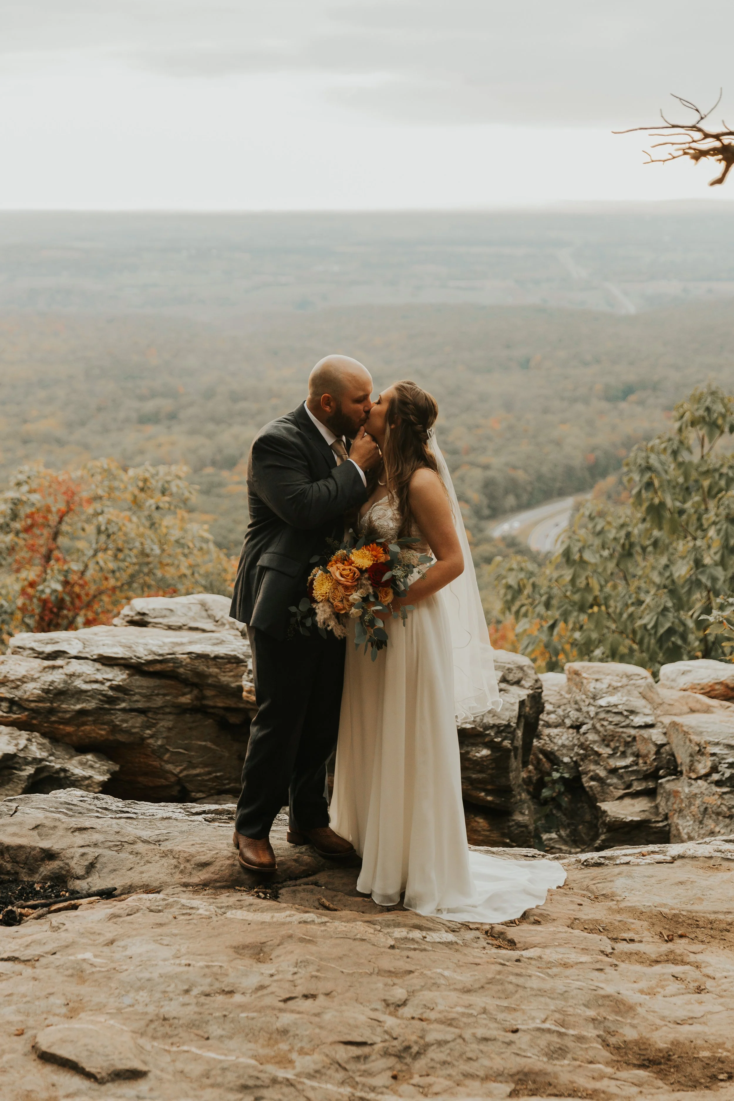 A couple is kissing during their wedding ceremony outdoors on a rocky outcrop with a scenic landscape of hills and trees in the background.