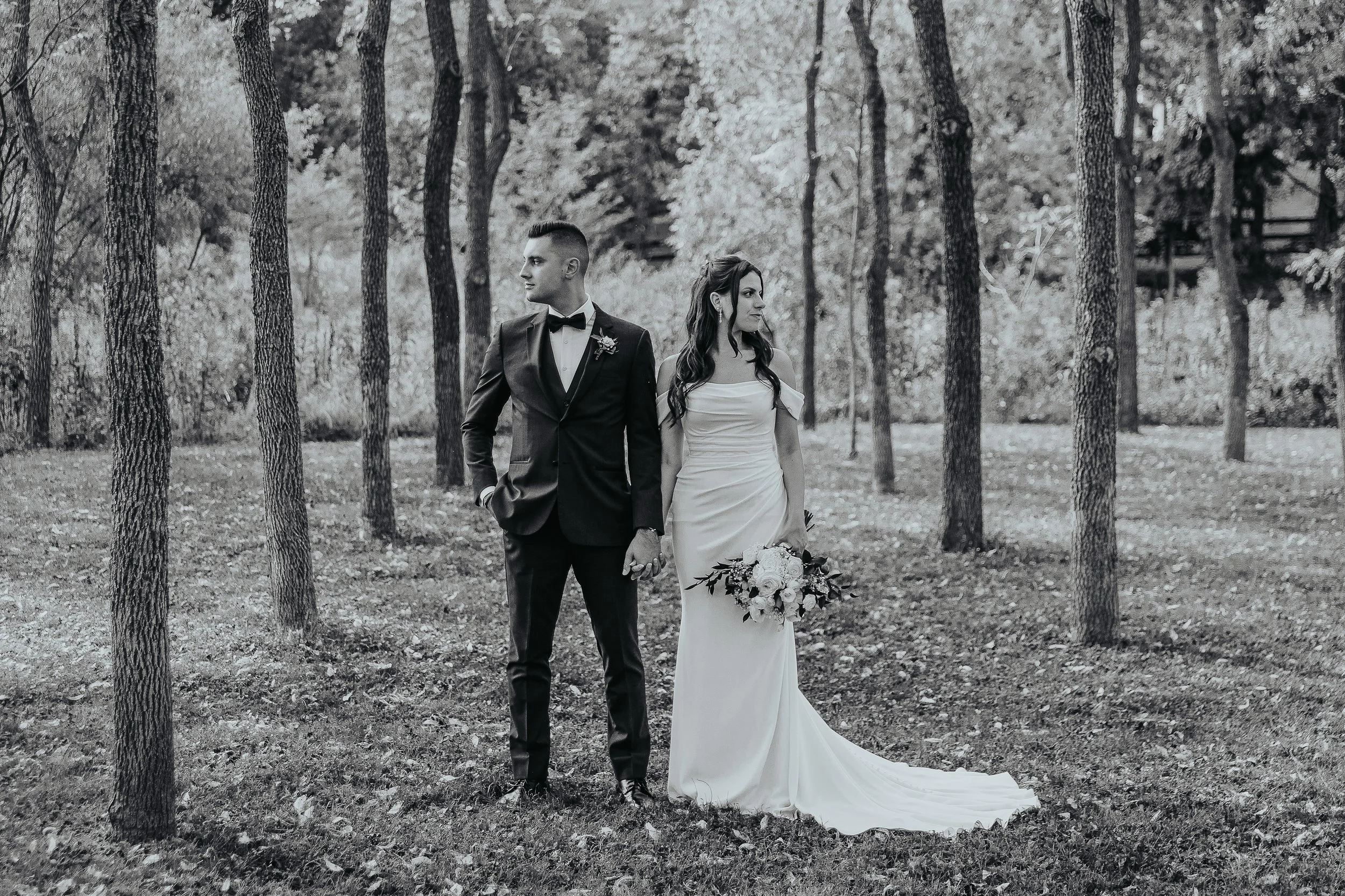 A black-and-white photo of a bride and groom holding hands outdoors among trees, the groom wearing a tuxedo and the bride in a strapless wedding dress holding a bouquet of flowers.