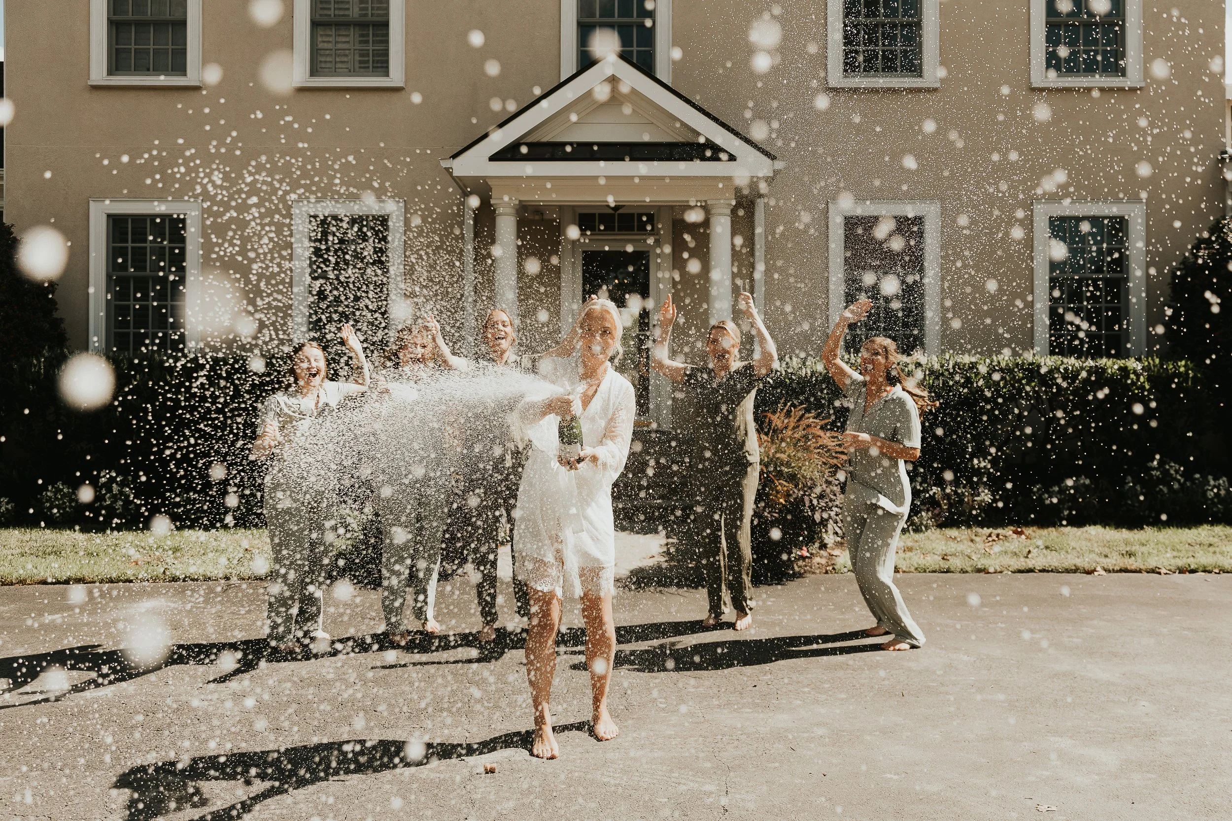 A group of people celebrating outside a house with a beige facade, white trim, and multiple windows. They are engaged in a water fight, with splashes of water and smiling faces. The scene suggests a joyful and fun gathering in warm weather.