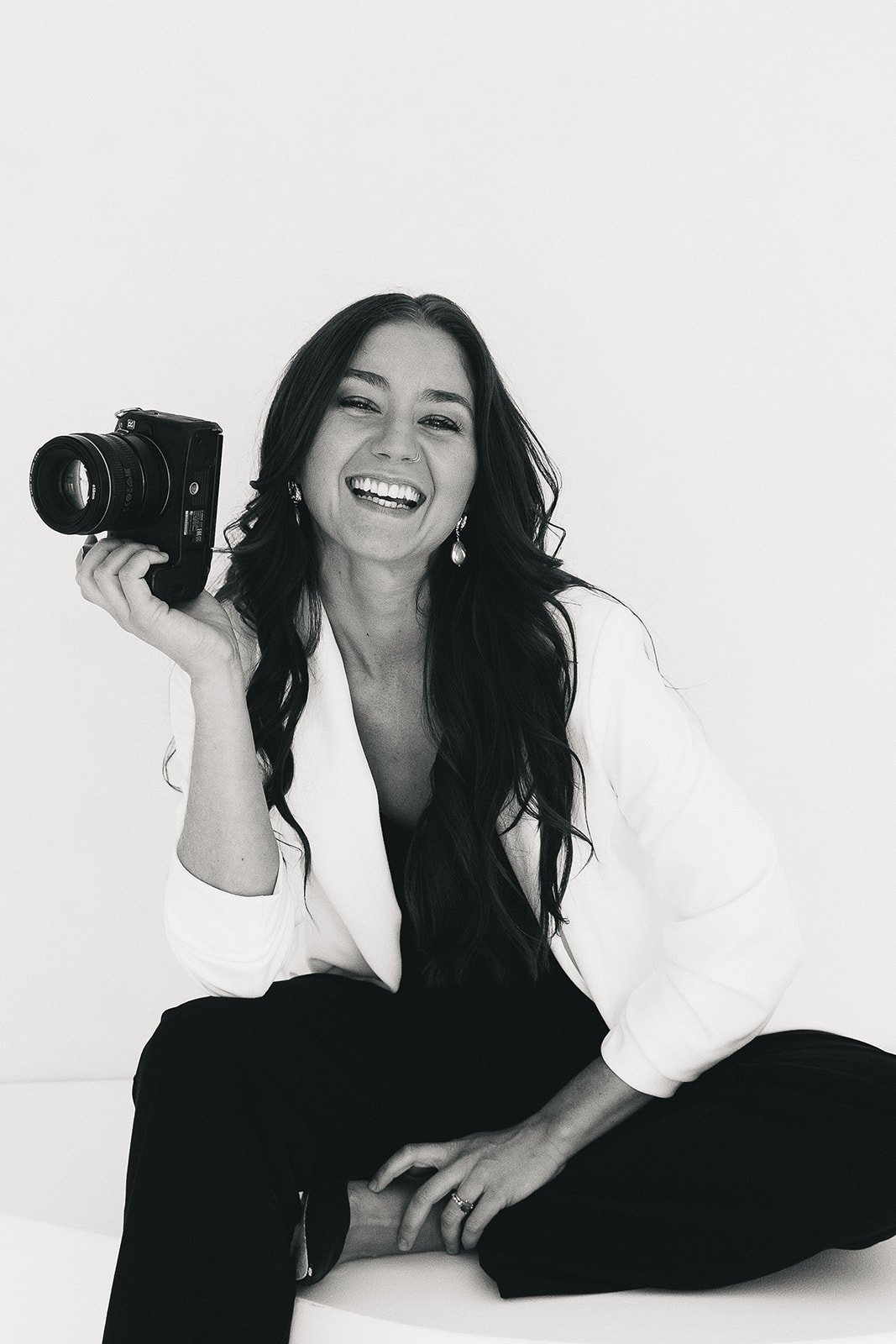 A woman with long dark hair, smiling, holding a camera in her right hand, dressed in a white blazer and black pants, sitting against a plain background.