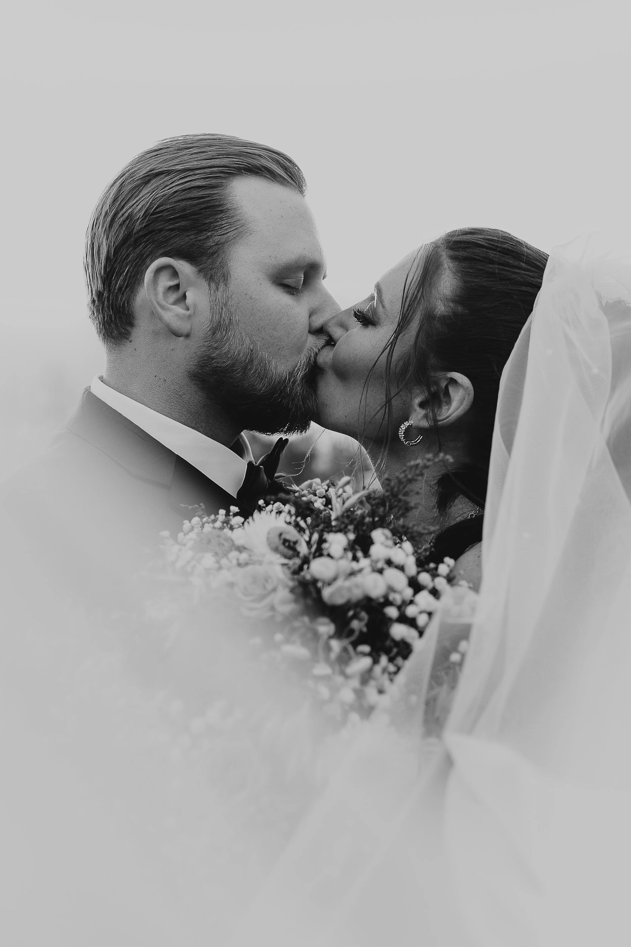 Black and white photo of a couple kissing, the man has a beard and closed eyes, the woman has her eyes open and is holding a bouquet of flowers, with a veil in the background.
