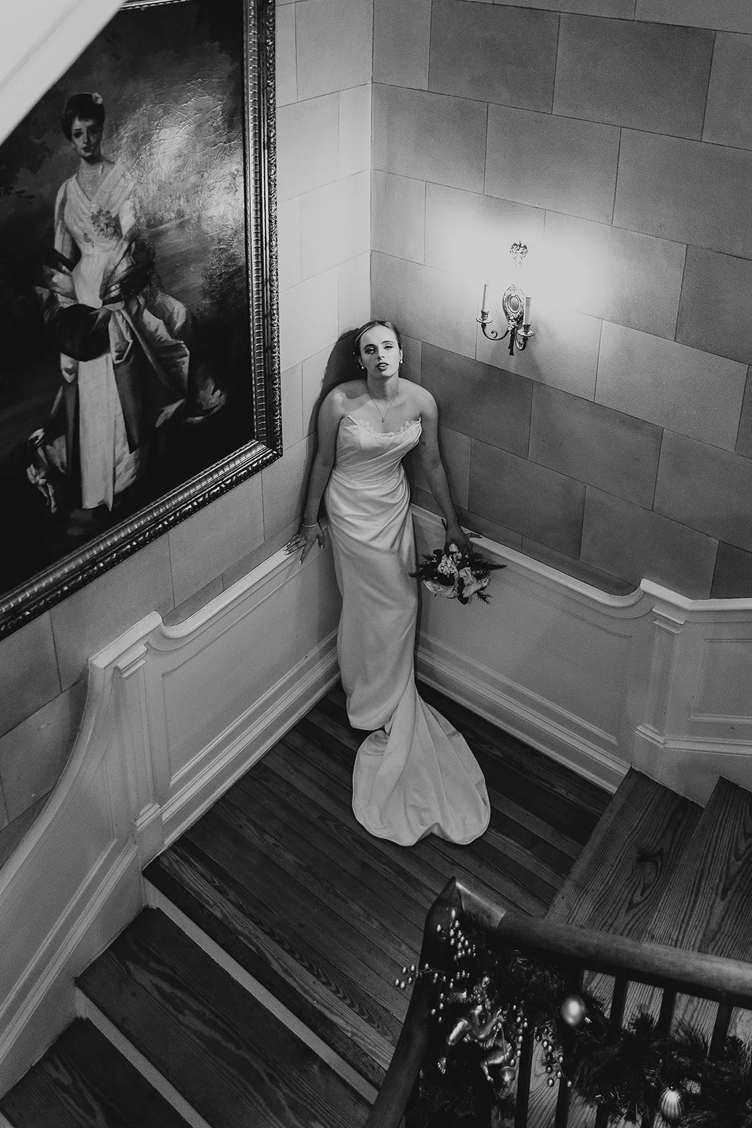A woman in a strapless wedding gown holding a bouquet, standing in a corner of a staircase landing with a large framed portrait on the wall next to her, illuminated by a wall-mounted lamp in a formal interior space.