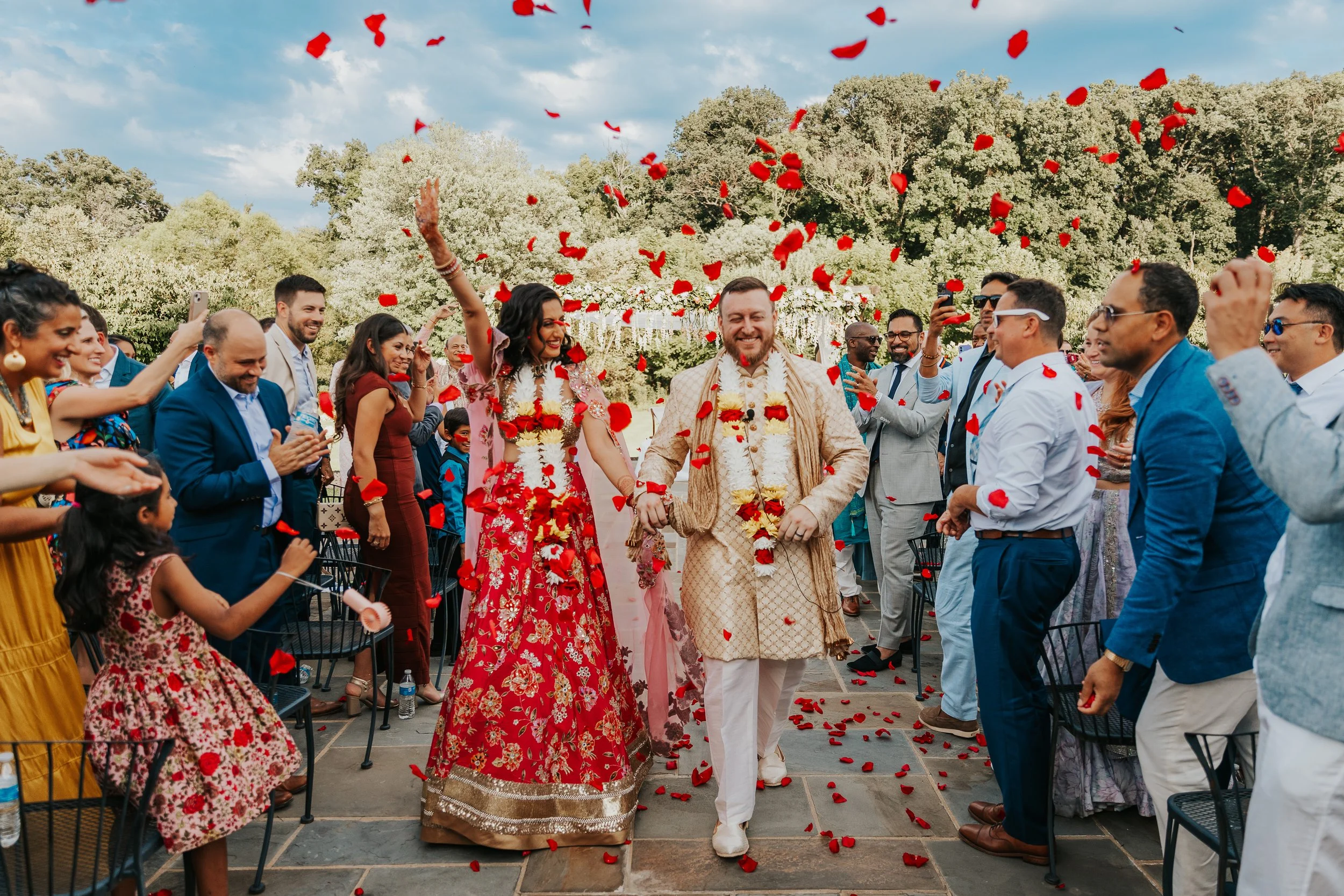 A wedding celebration outdoors with a bride and groom walking through a flower petal shower surrounded by guests smiling and taking photos.