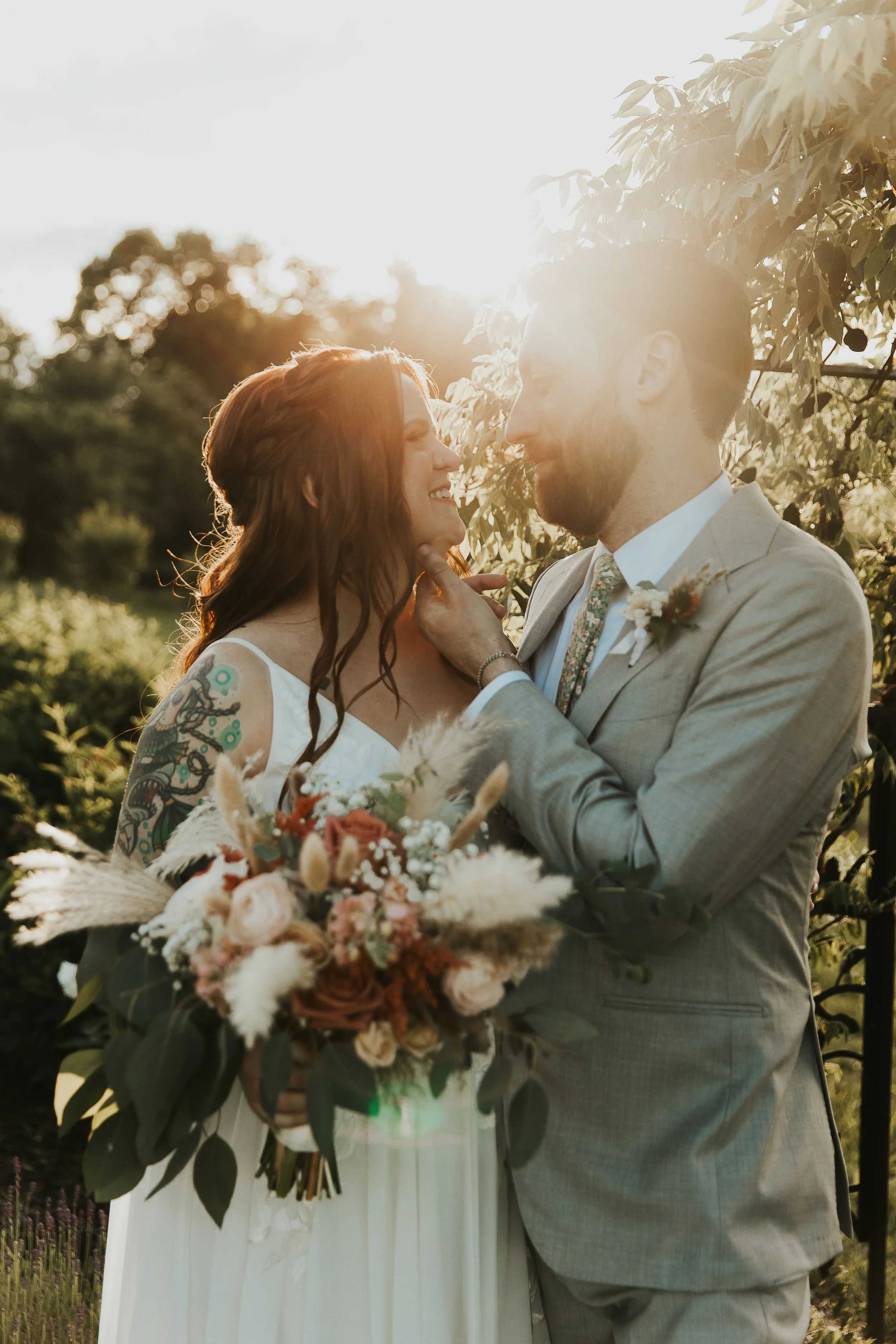 An elopement couple in wedding attire, holding a bouquet, standing outdoors in a garden with green foliage, during sunset, sharing a joyful moment.