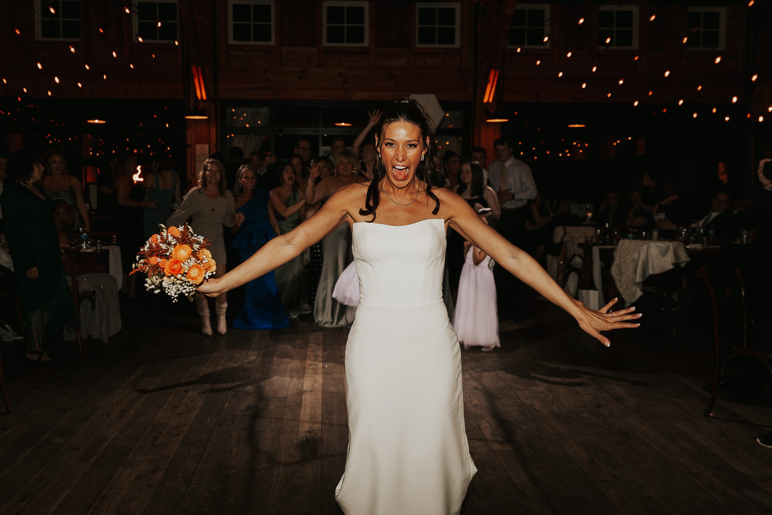 A woman in a white wedding dress with dark hair styled in curls, smiling and dancing at a wedding reception with guests in the background under warm, festive lighting.