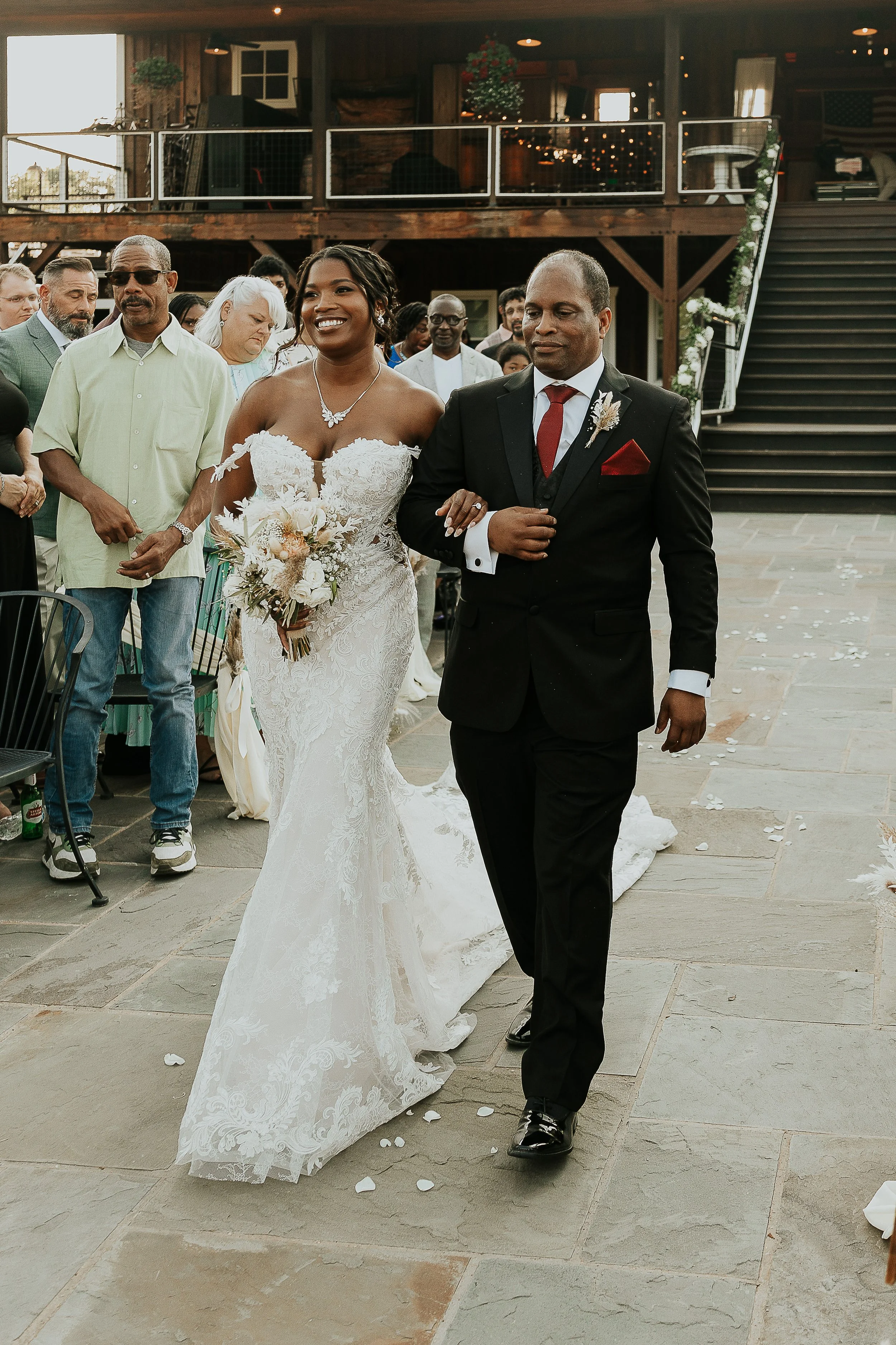 A bride and groom walking arm in arm during their wedding ceremony, surrounded by guests, on a stone patio with scattered flower petals.
