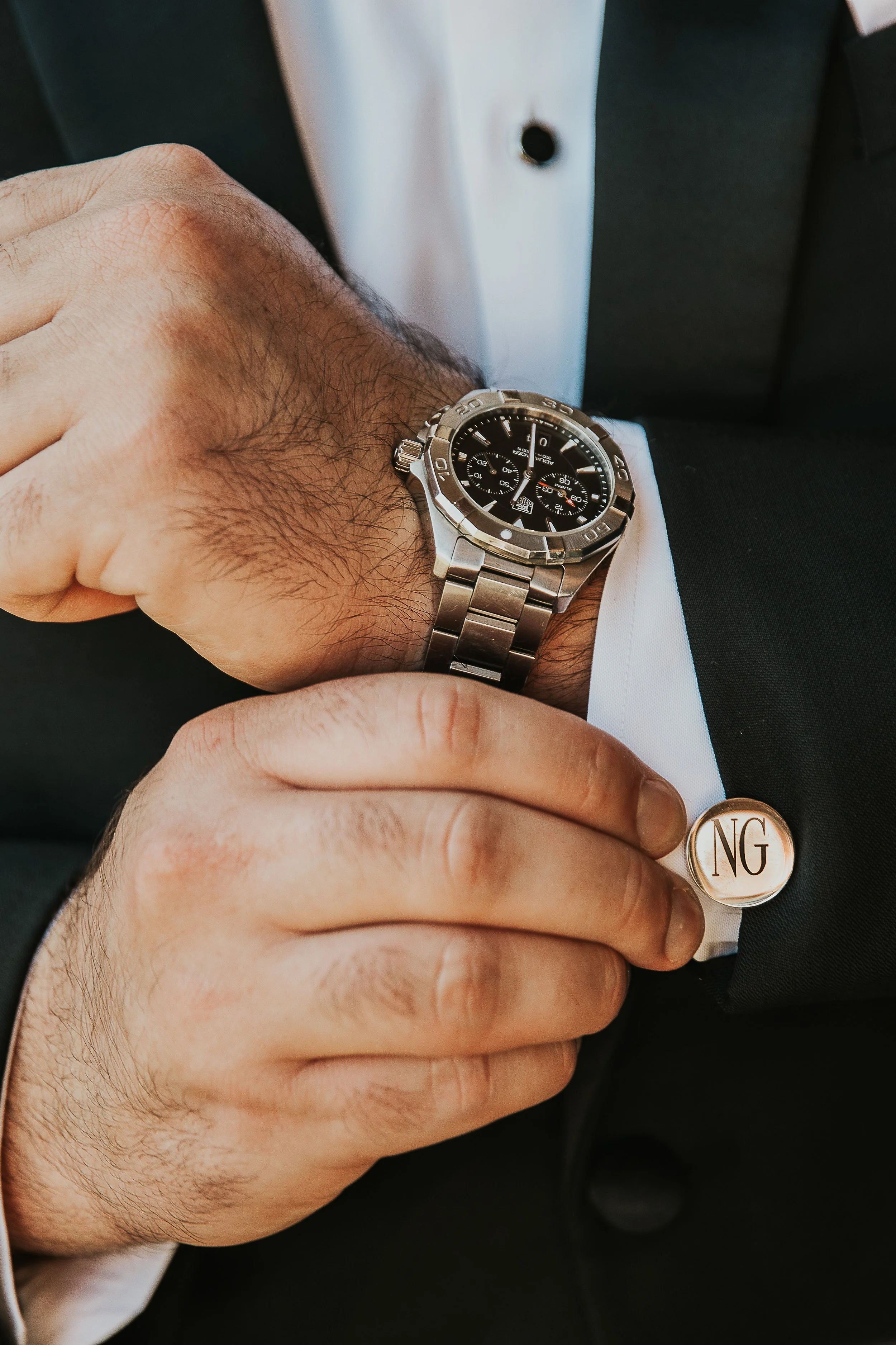 A person wearing a silver wristwatch with a black dial and a chronograph, adjusting a cufflink on a white shirt sleeve. The cufflink has gold initials 'NG' on it. The person is dressed in a black suit.