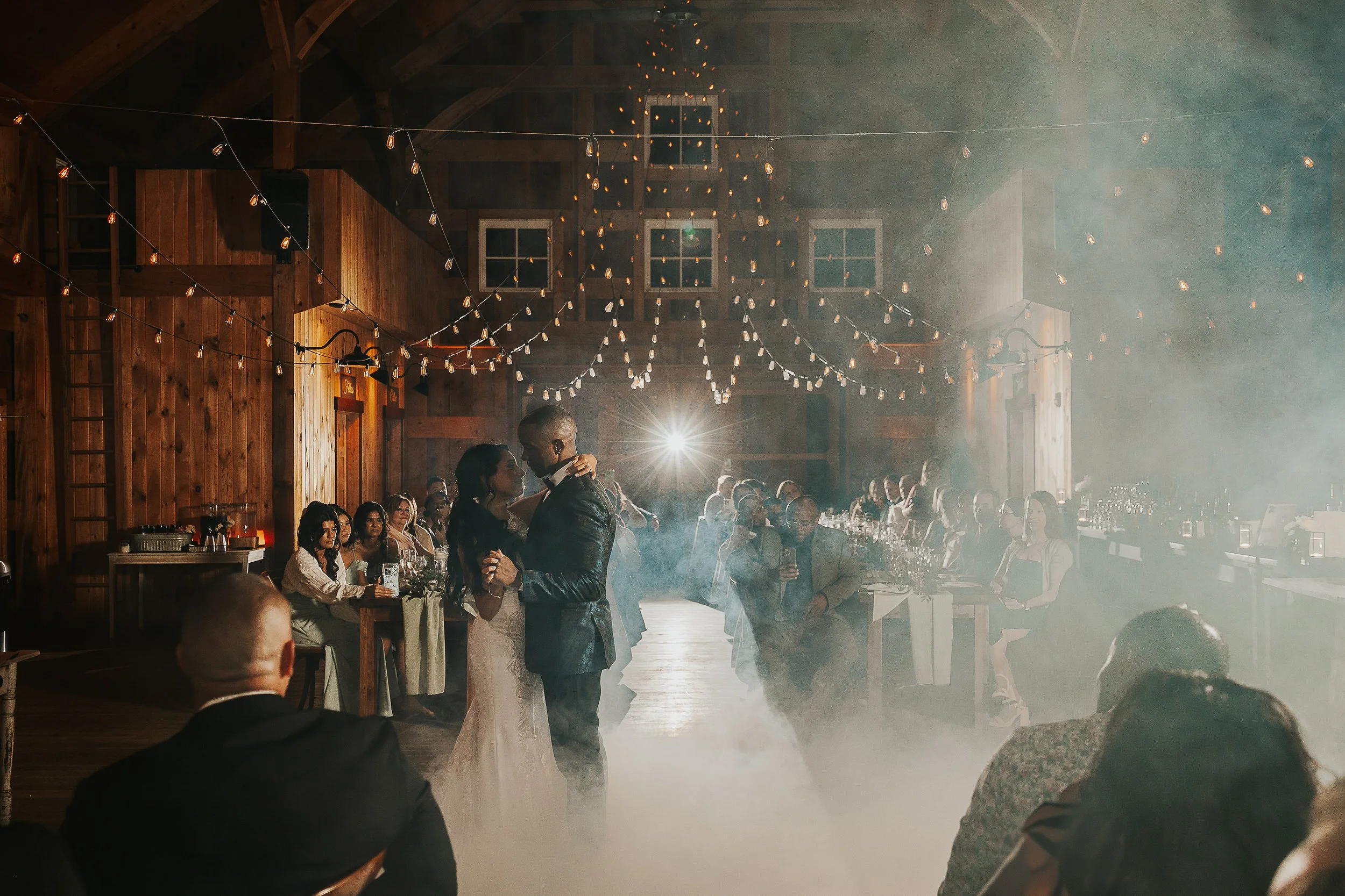 A bride and groom are dancing in the center of a rustic wedding reception held in a barn with wooden walls and string lights hanging from the ceiling. Guests are seated around tables, watching the couple, with some taking photos. The atmosphere is wa