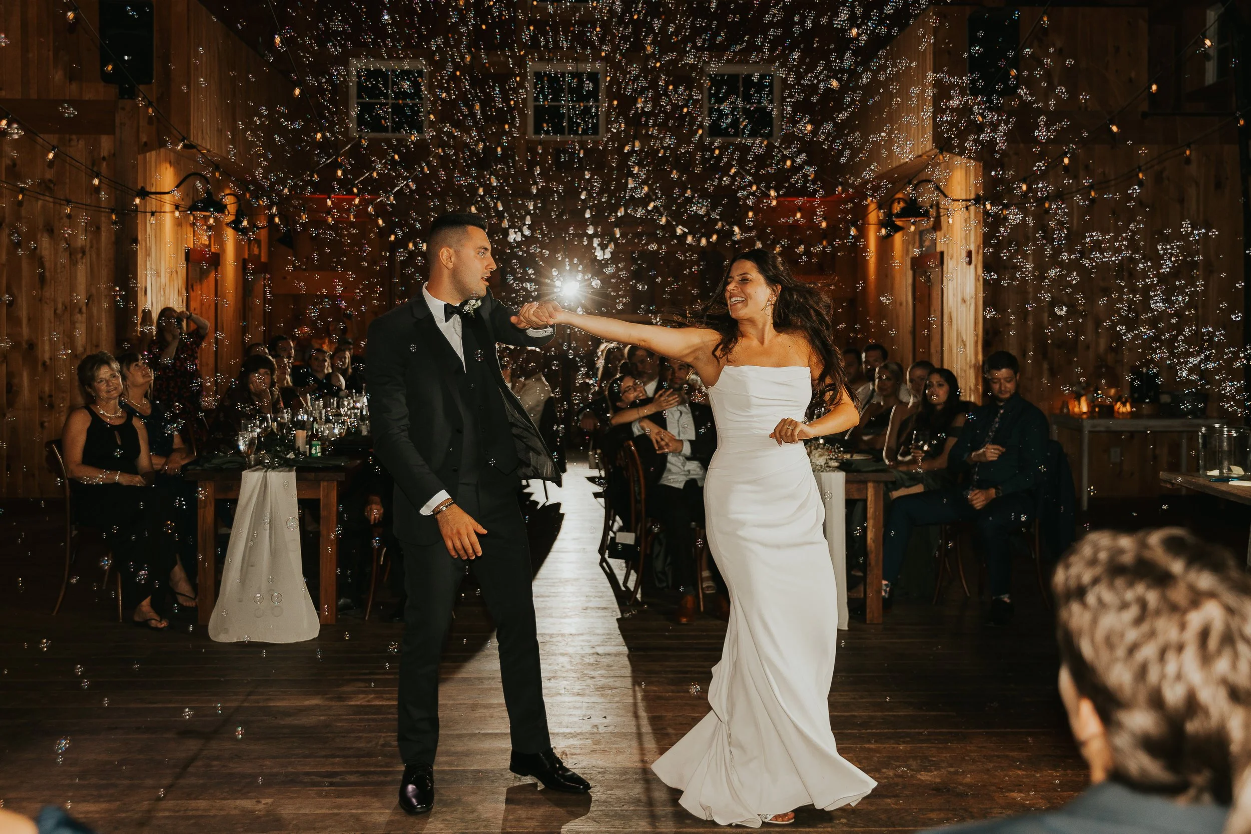 A bride and groom dancing at a wedding reception under a ceiling decorated with string lights and bubbles, with seated guests watching.