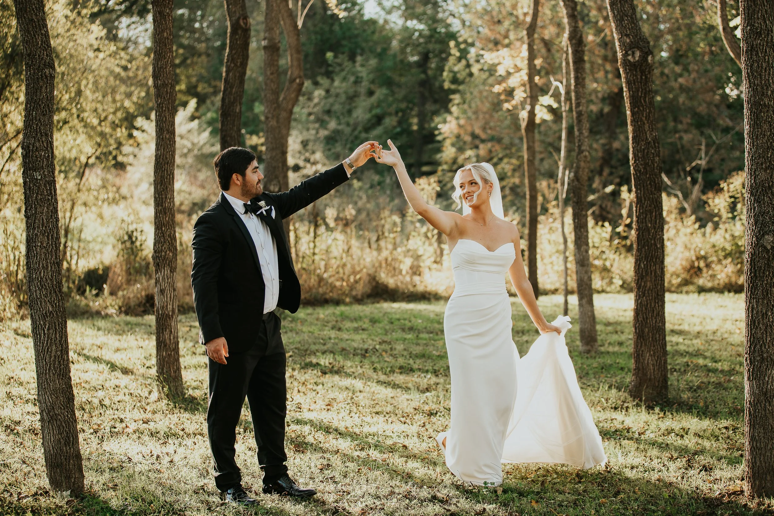 A bride and groom dancing or celebrating outdoors in a wooded area during daylight, with trees and green grass surrounding them.