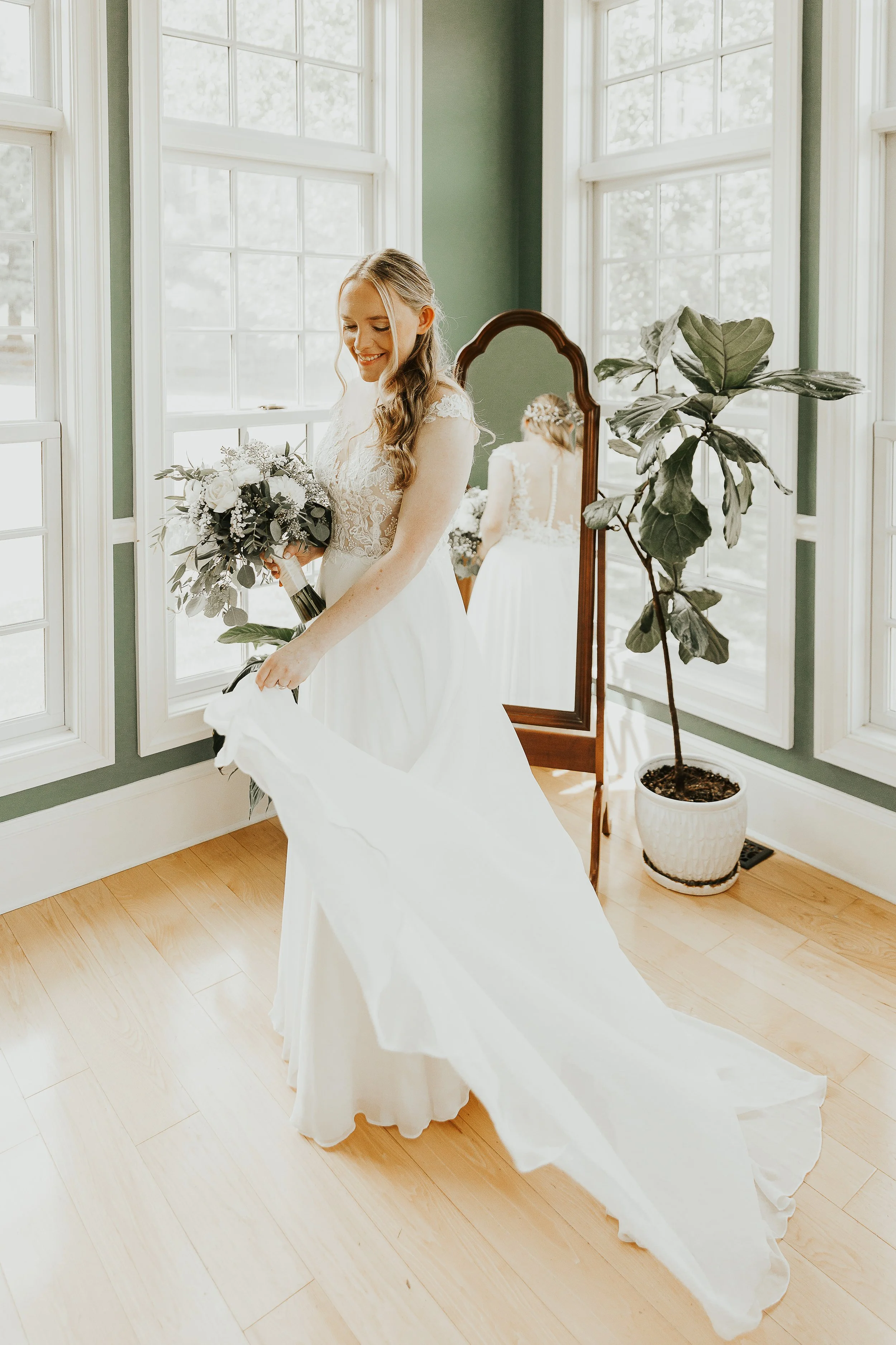 Bride in a white wedding dress holding a bouquet, smiling while looking down, with an elegant mirror and green potted plant in a sunlit room with large windows.