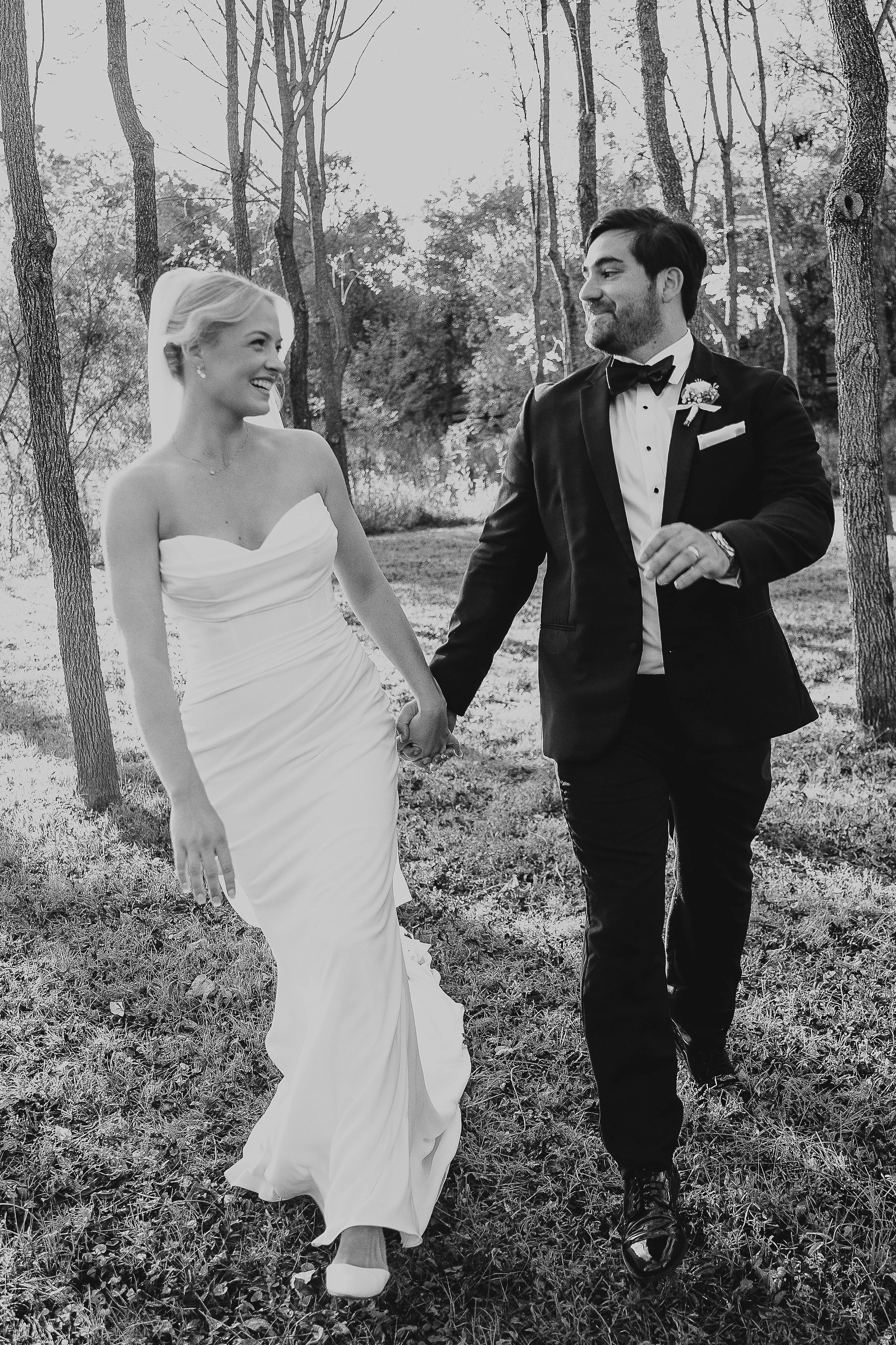 Black and white photo of a bride and groom outdoors in a wooded area, walking hand in hand. The bride is wearing a strapless wedding gown and the groom is in a tuxedo with a bow tie, both smiling at each other.