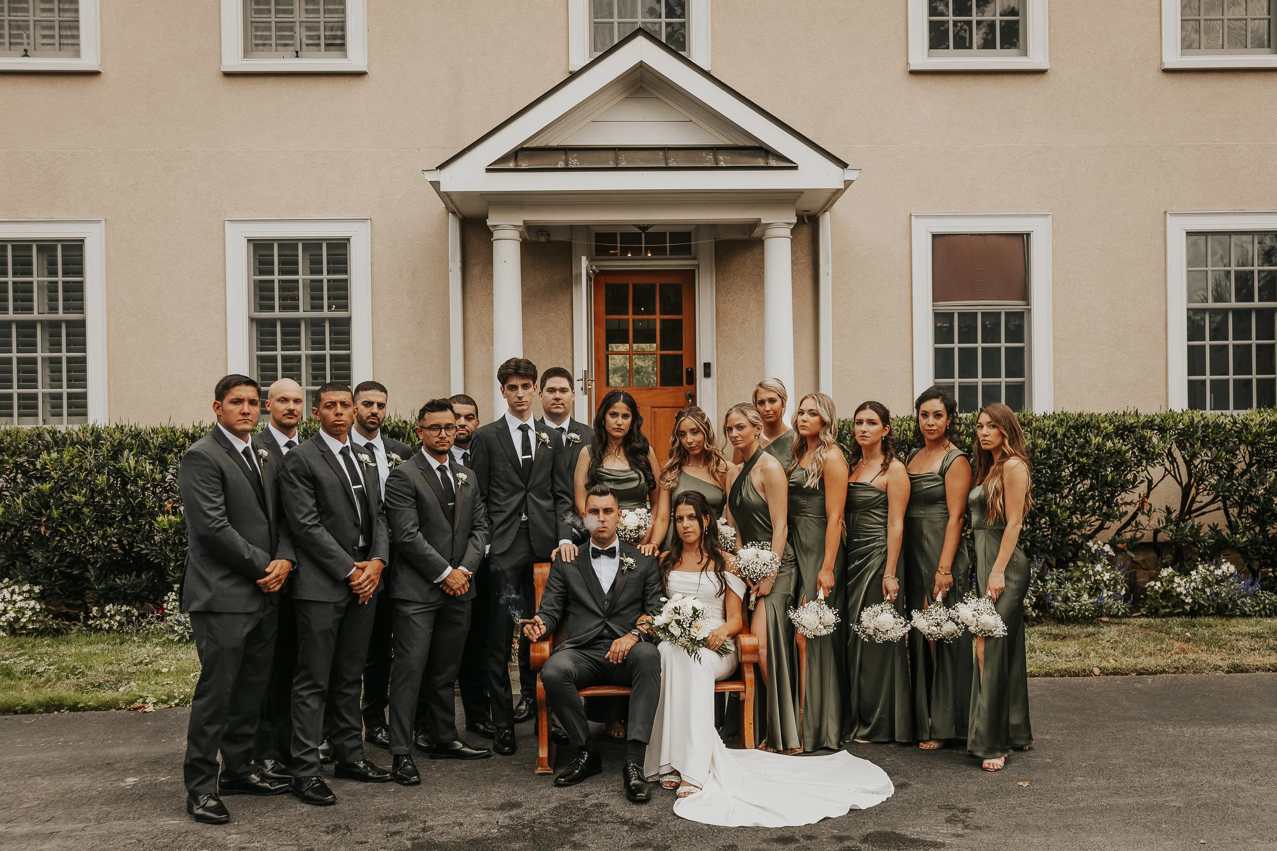 A large group of wedding guests standing outside in front of a beige house with white trim and multiple windows. The bride and groom are seated in front, surrounded by bridesmaids and groomsmen. The men are dressed in suits with ties, and the women a