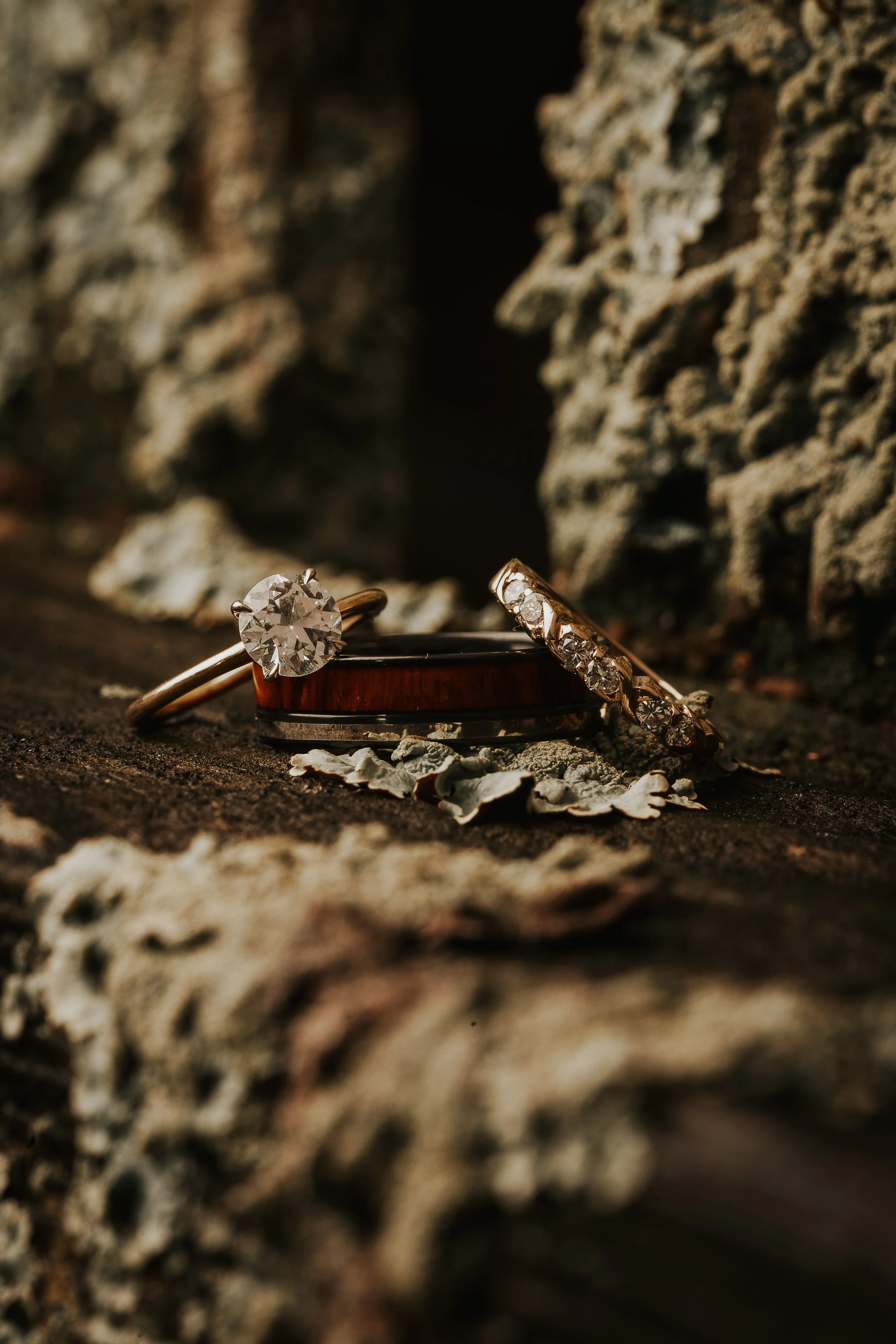 Close-up of three rings, two with diamonds and one with smaller stones, placed on a rough, textured surface with rocks surrounding them.