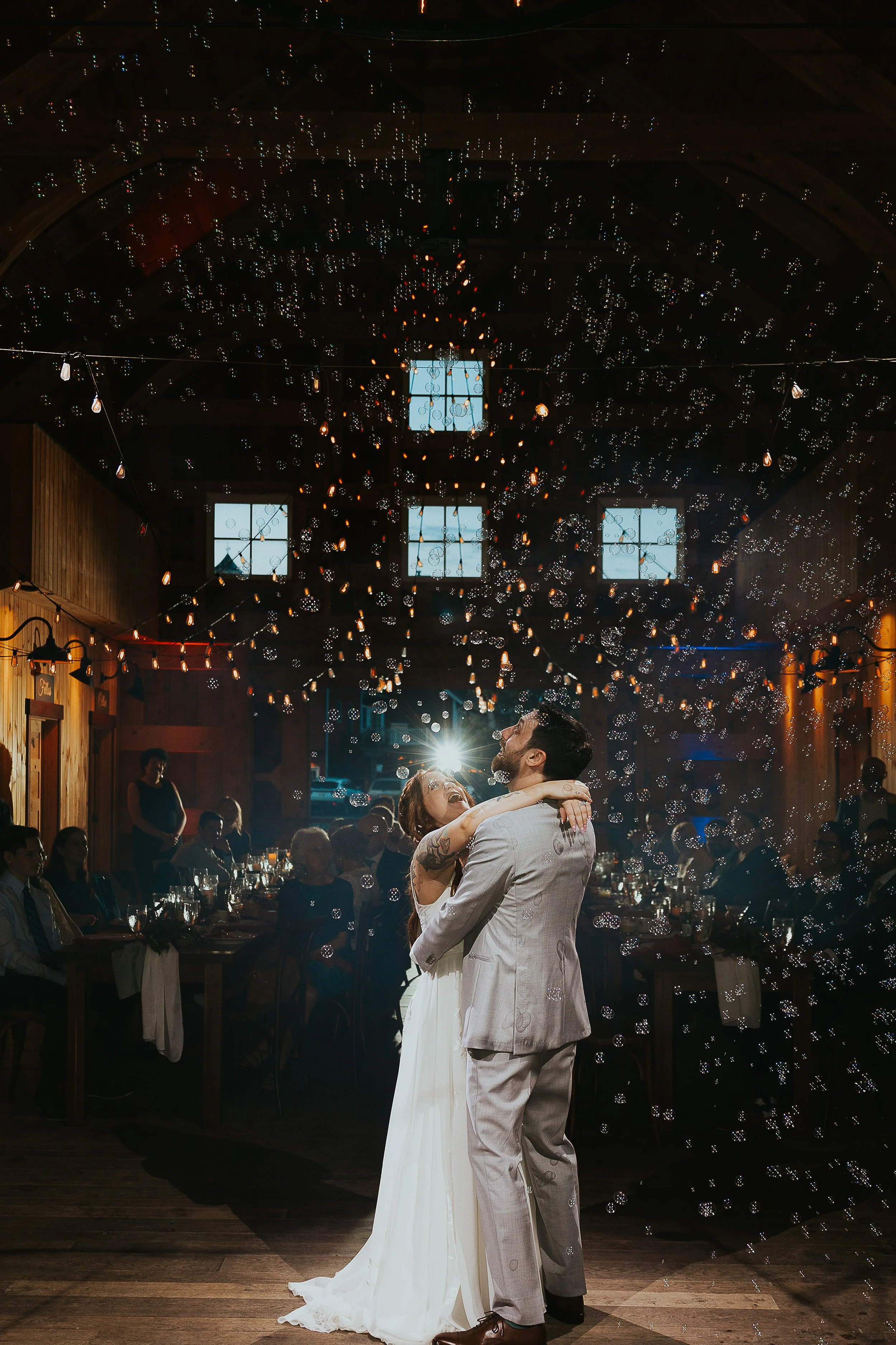 A newlywed couple dancing at their wedding reception, surrounded by guests in a barn decorated with string lights and bubbles, with natural wood walls and high windows.