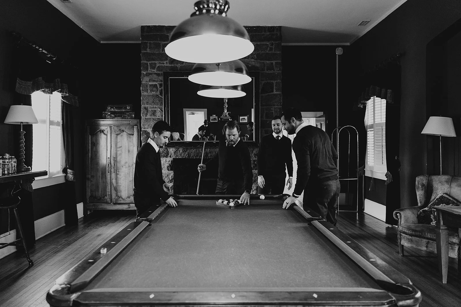 Four men are playing billiards in a cozy, dimly lit room with a stone fireplace, wooden furniture, and curtains on the window.