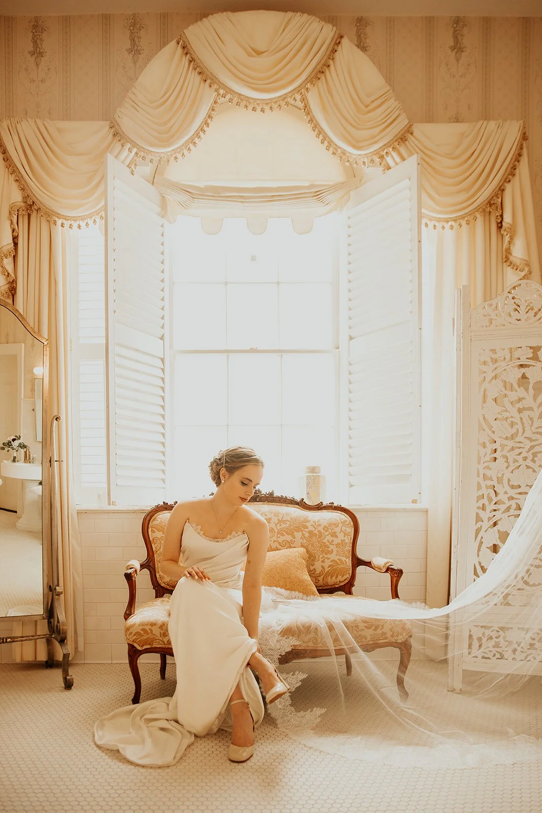 A woman in a white wedding dress sits on an ornate vintage sofa in a bright room with large windows, white blinds, and elegant curtains.