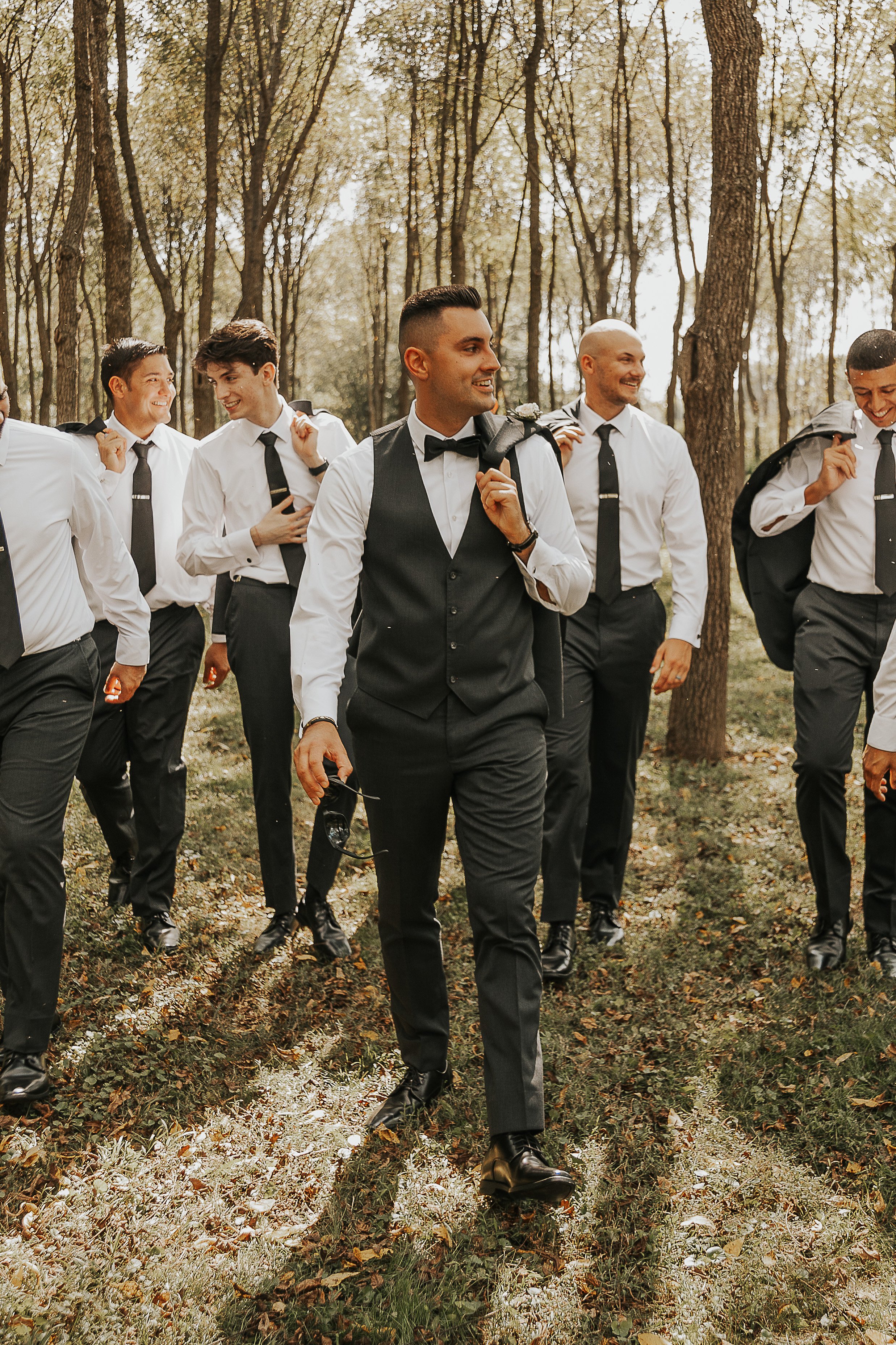 Group of men dressed in formal wear with white shirts, black ties, and black vests or jackets walking in a forested area during daytime.