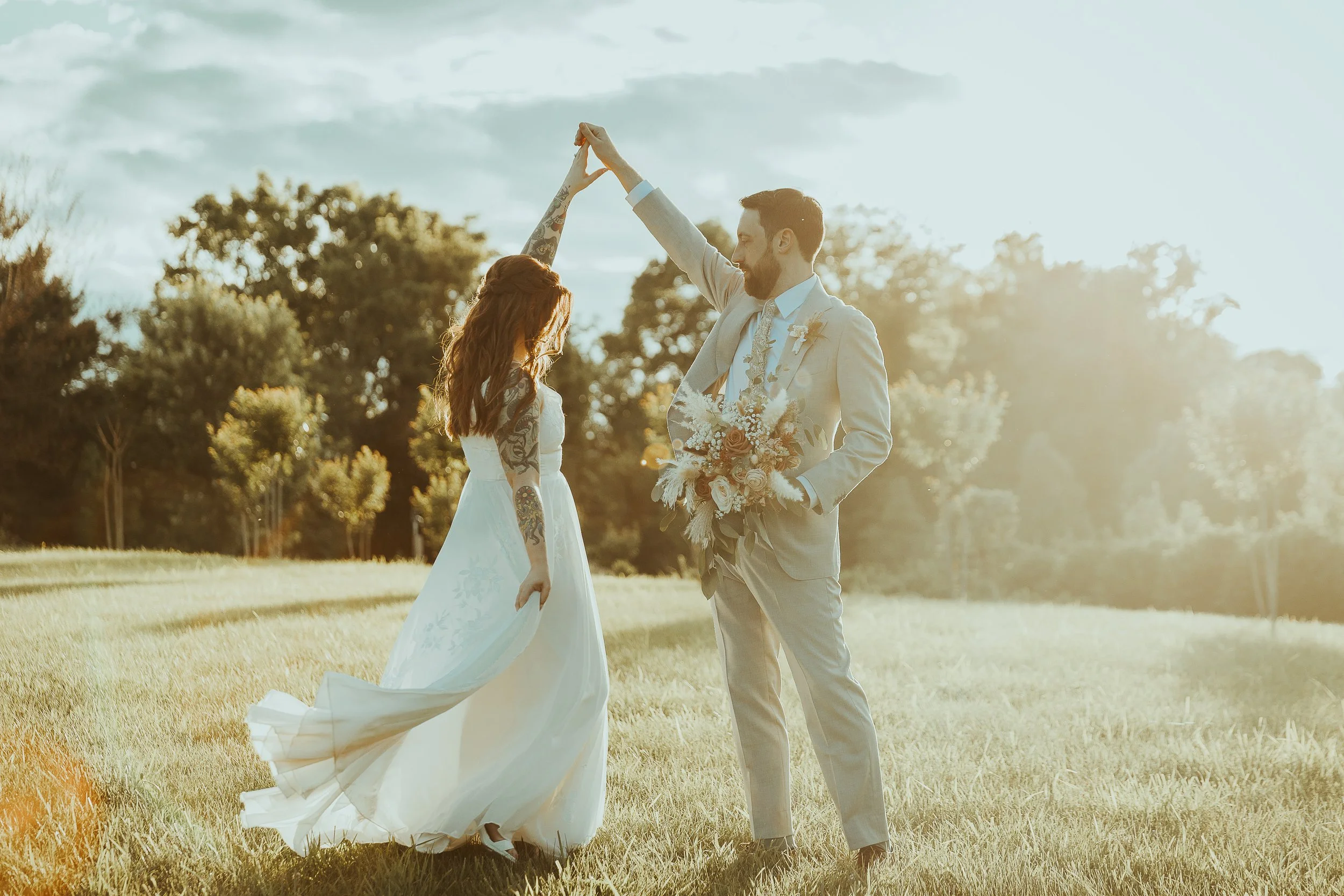 A couple dressed in wedding attire dancing outdoors during sunset, with trees and a cloudy sky in the background.