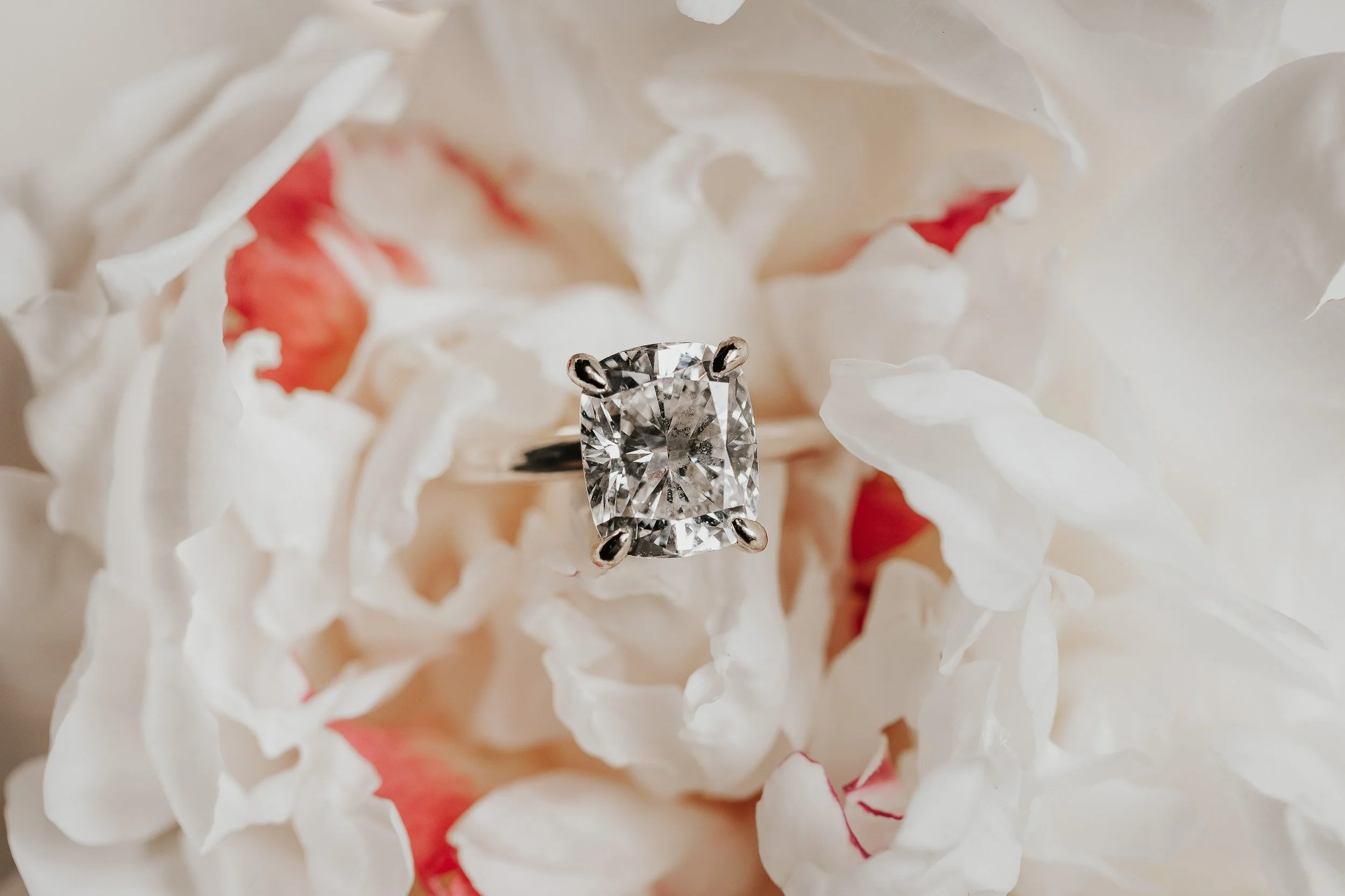 A close-up image of a diamond ring with a large, square-cut diamond held in prongs, placed on white and pink flower petals.