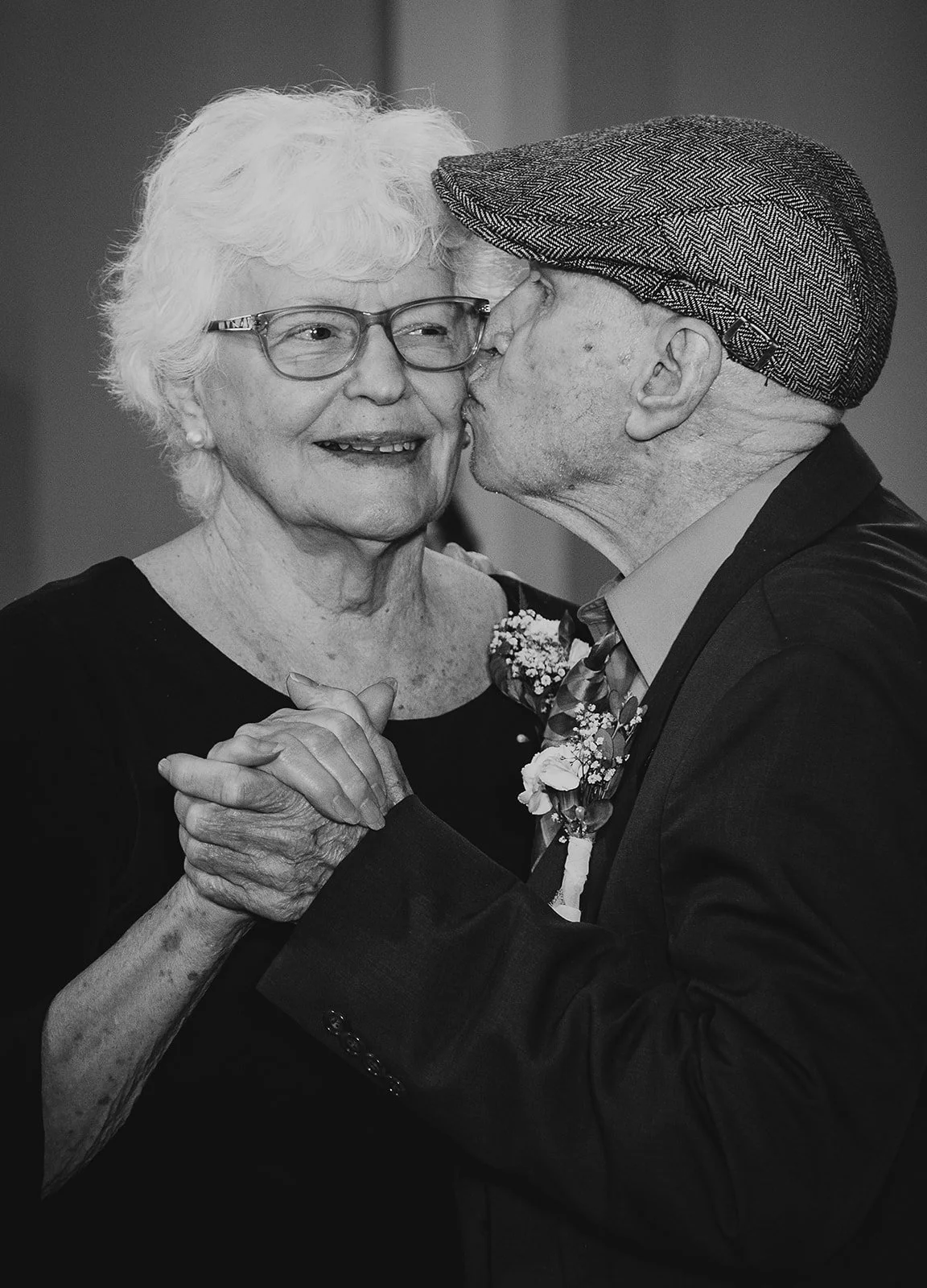 An elderly woman with glasses smiling as an elderly man, wearing a hat, kisses her on the cheek during a dance.