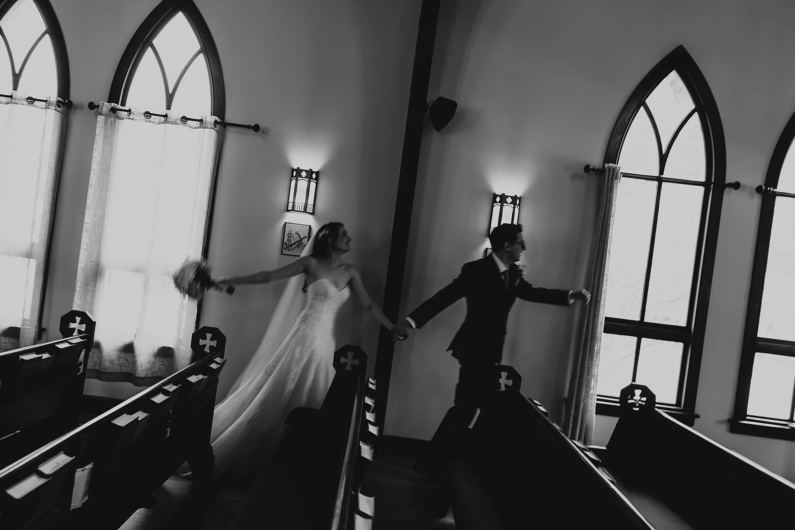 Black and white photo of a bride and groom walking hand in hand inside a church, with church pews and large arched windows in the background.