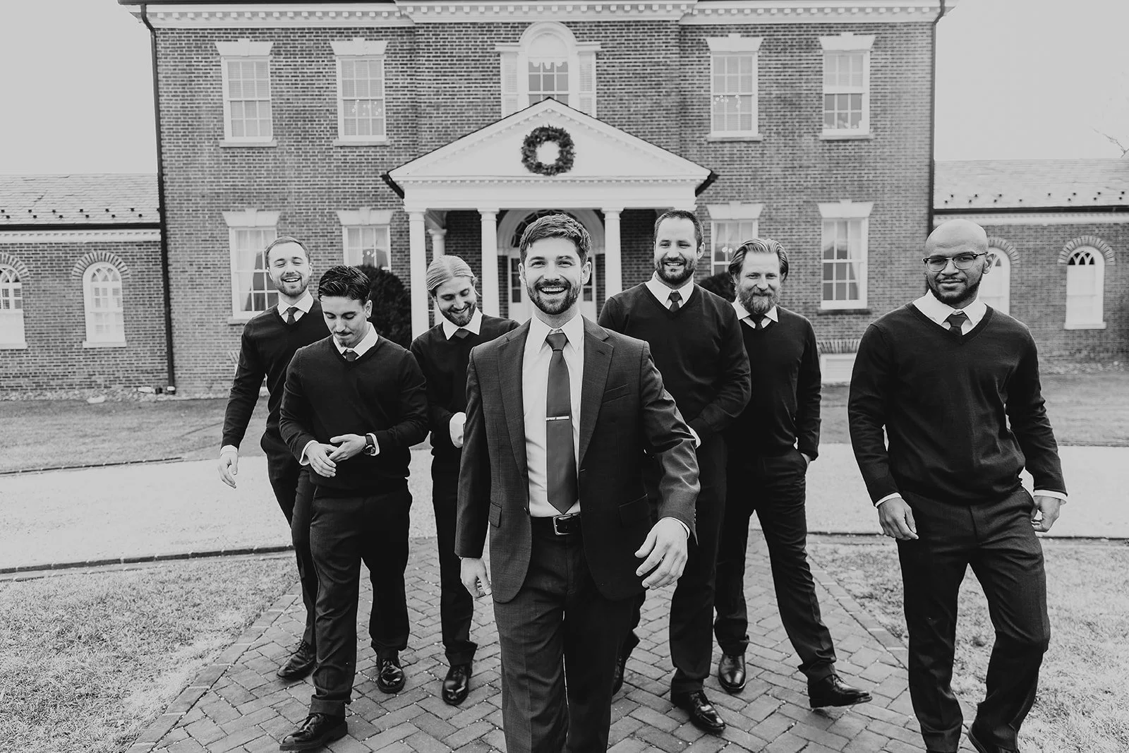 A group of seven men dressed in suits and sweaters walking outside on a paved pathway in front of a large brick house with a wreath hanging on the front. The men are smiling and appear to be having a good time.
