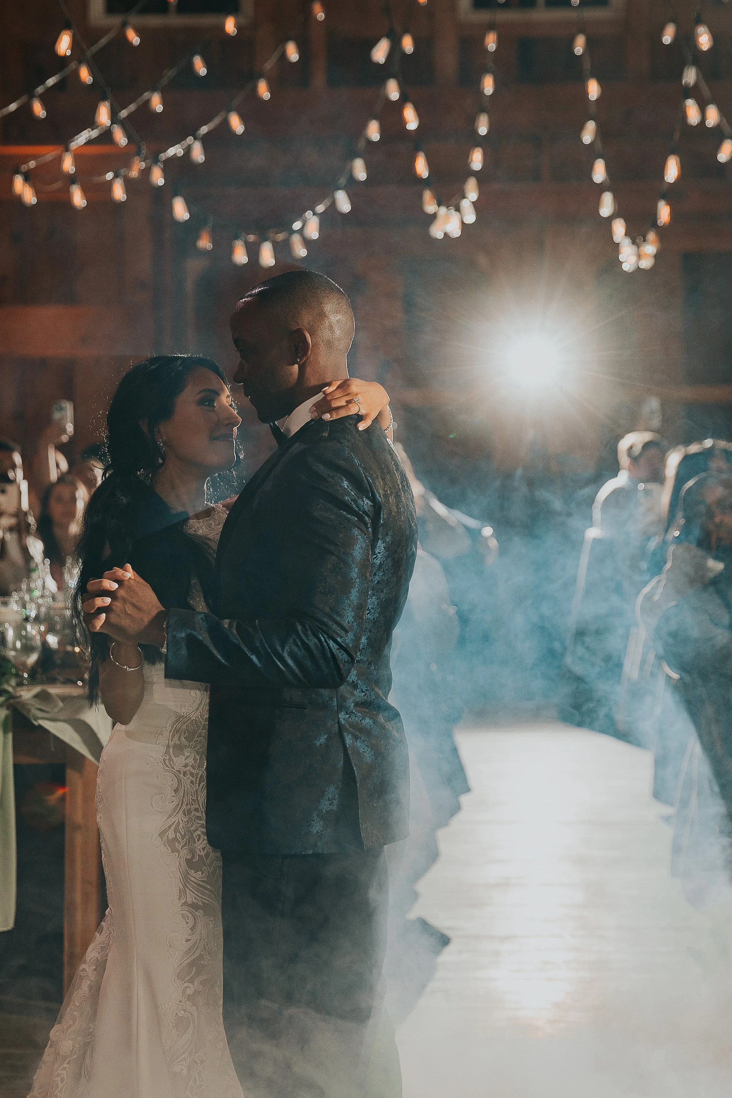 A couple dancing closely during their wedding reception, with wedding guests seated at tables in the background and string lights hanging from the ceiling.
