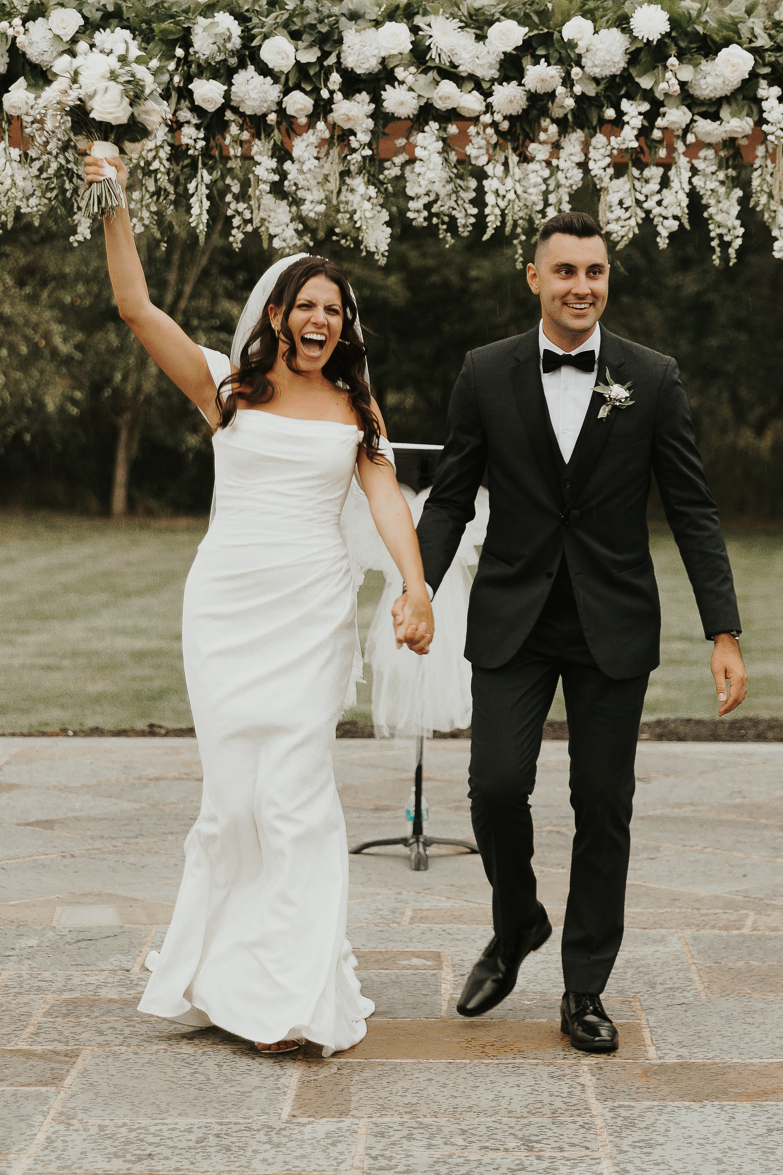 A bride and groom are holding hands and walking outdoors during their wedding. The bride is cheering with her mouth open, holding a bouquet, and wearing a white wedding dress. The groom is smiling, dressed in a black tuxedo with a bow tie, and has a 