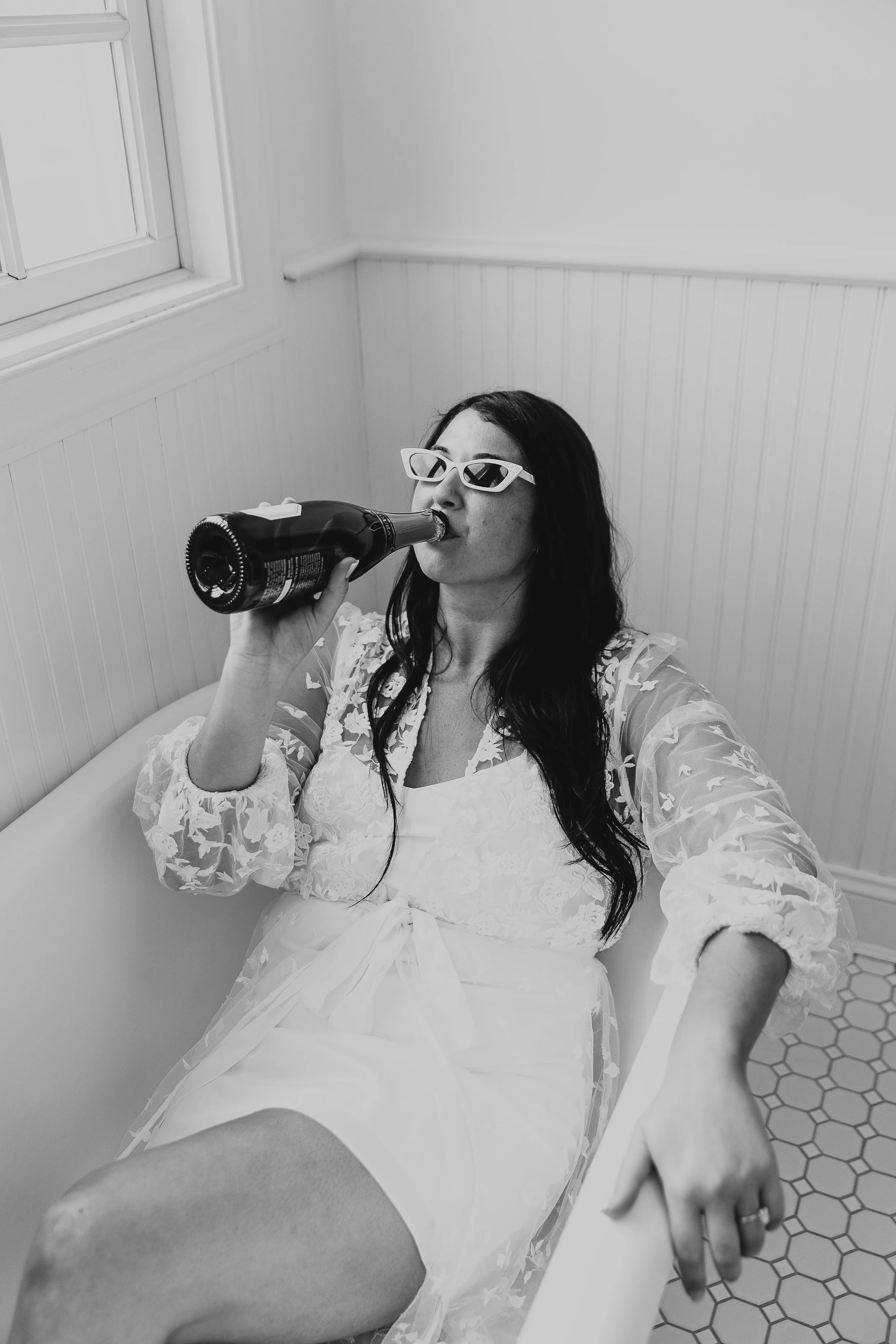 A woman with long dark hair wearing sunglasses and a white lace dress, sitting on a bench, drinking from a bottle.