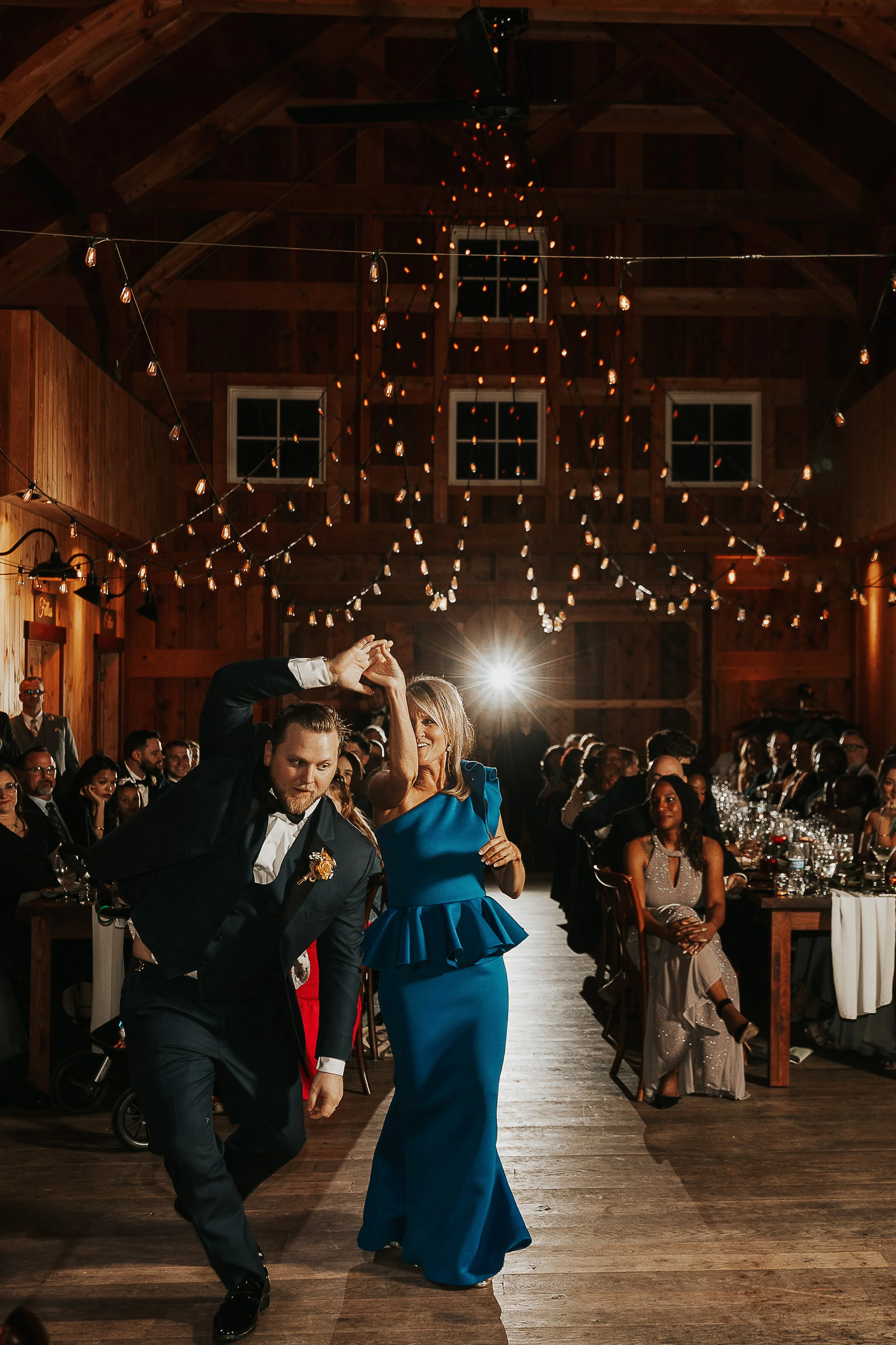 A man and a woman dancing together at a wedding reception in a rustic barn, with other guests seated at tables in the background and string lights hanging from the ceiling.