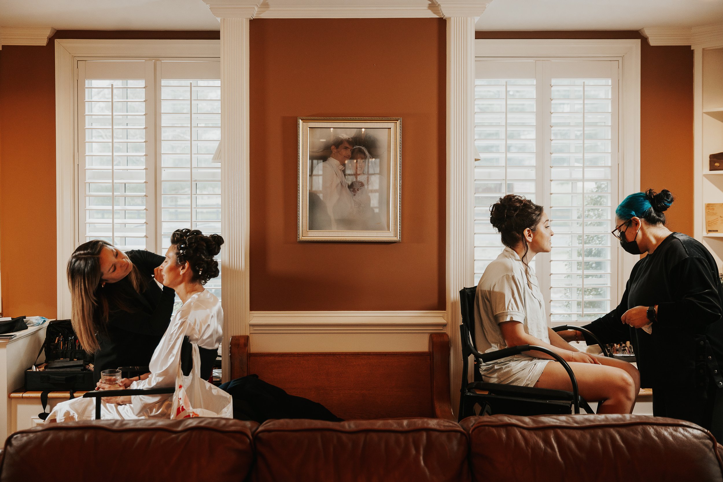 People preparing makeup and hairstyling for a bride in a room with two large windows and a framed picture on a brown wall.