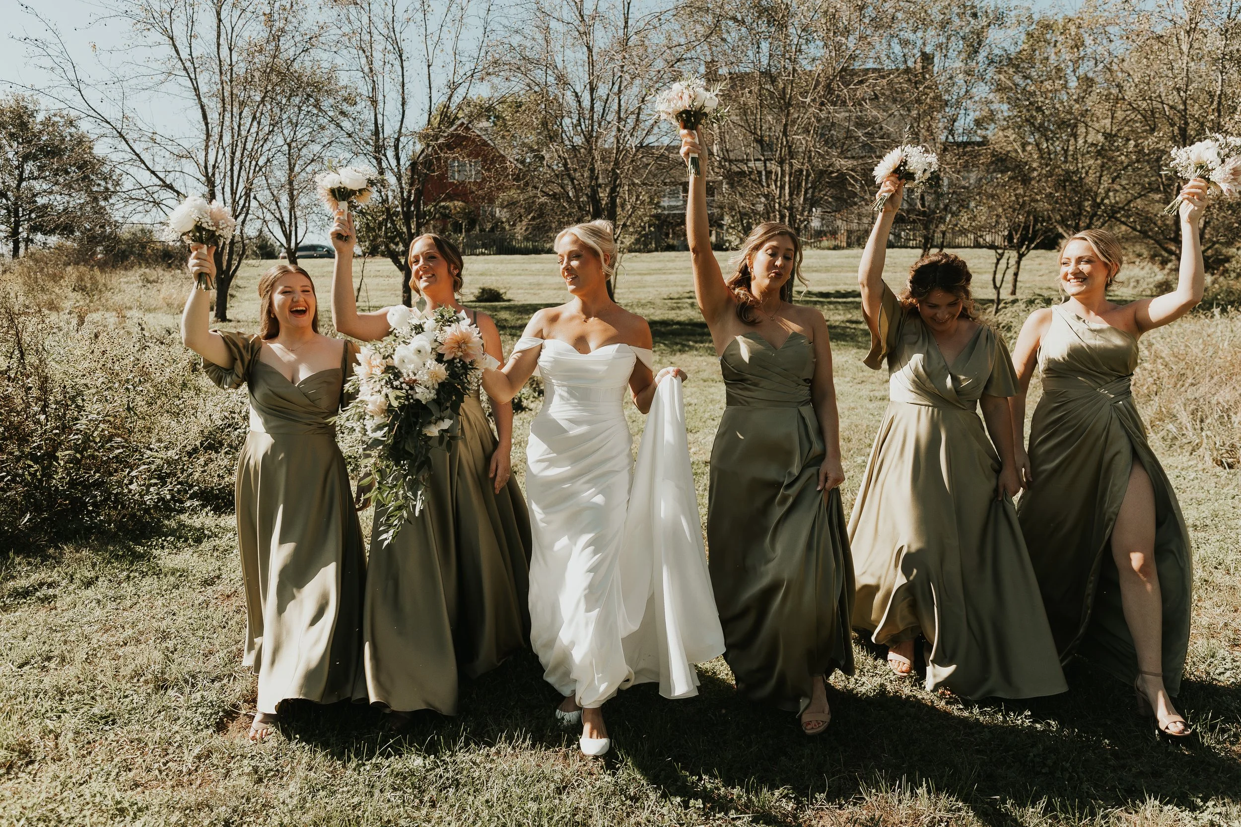 Group of bridesmaids and a bride walking outdoors on a sunny day, holding bouquets and celebrating.