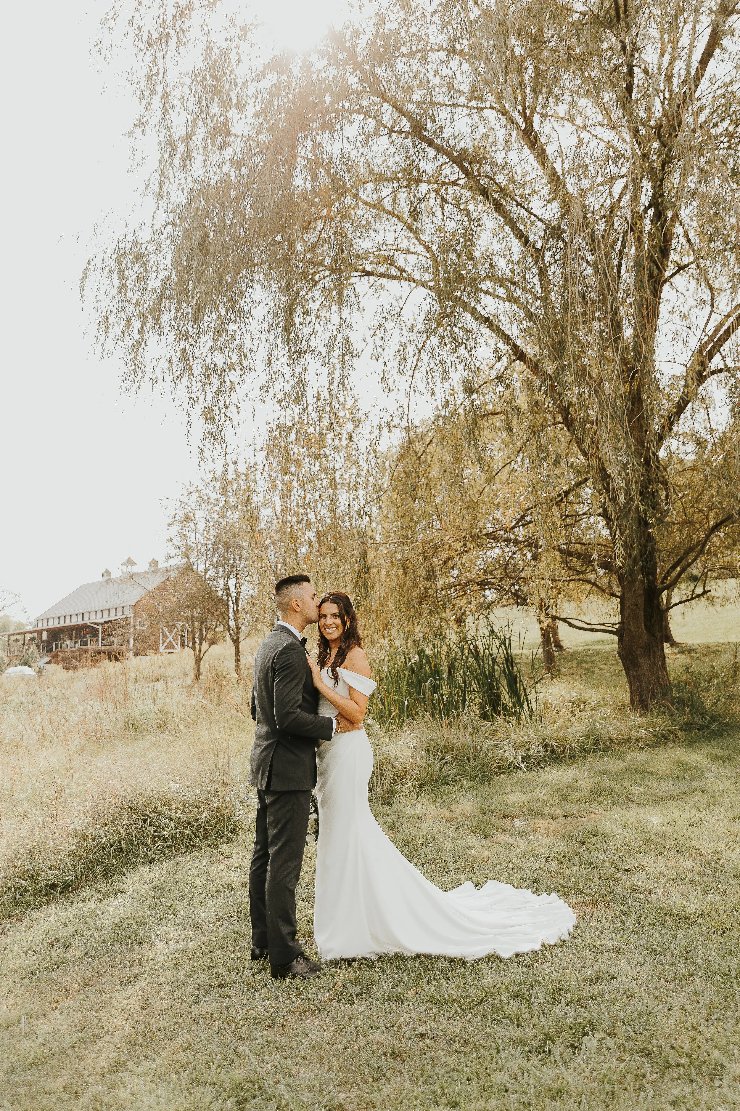 A couple in wedding attire standing outdoors under a large tree, the groom kissing the bride on her cheek, with a barn-like building and open field in the background, during daytime.