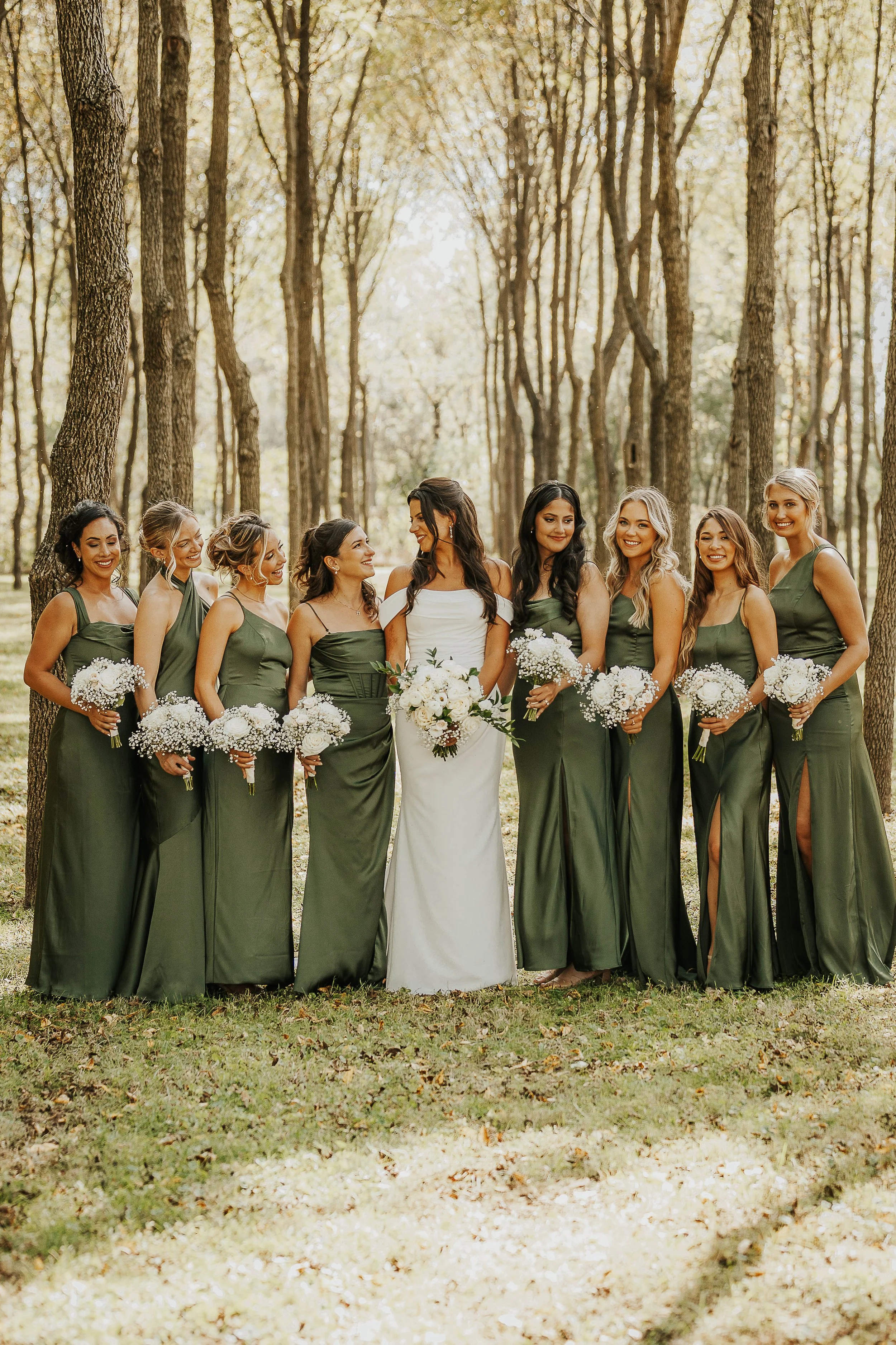 A group of women in bridesmaid dresses and a bride in a wedding gown standing together outdoors in a wooded area, holding bouquets of white flowers.