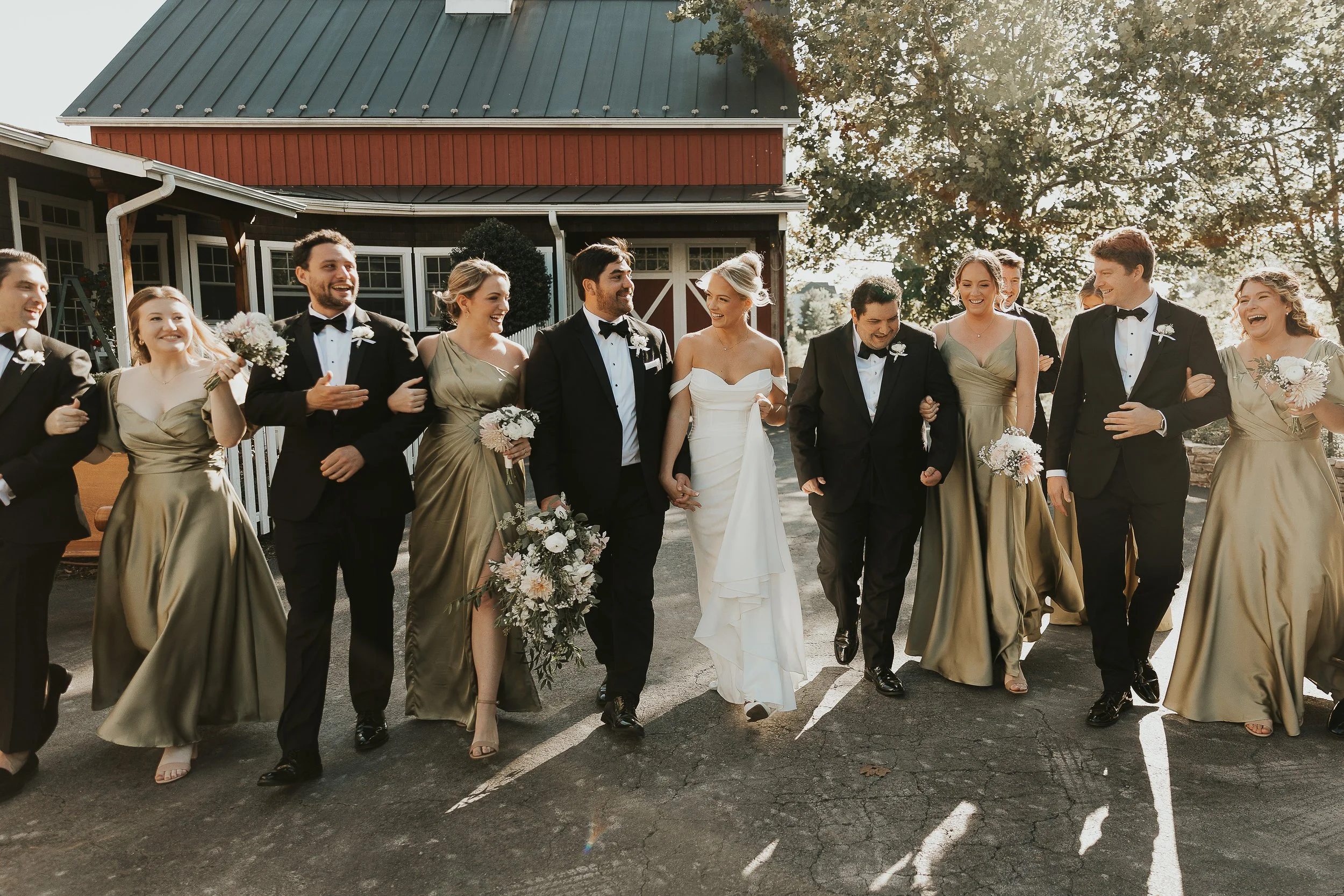 Group of people dressed in formal wedding attire walking arm in arm outside in front of a red barn, smiling and celebrating.