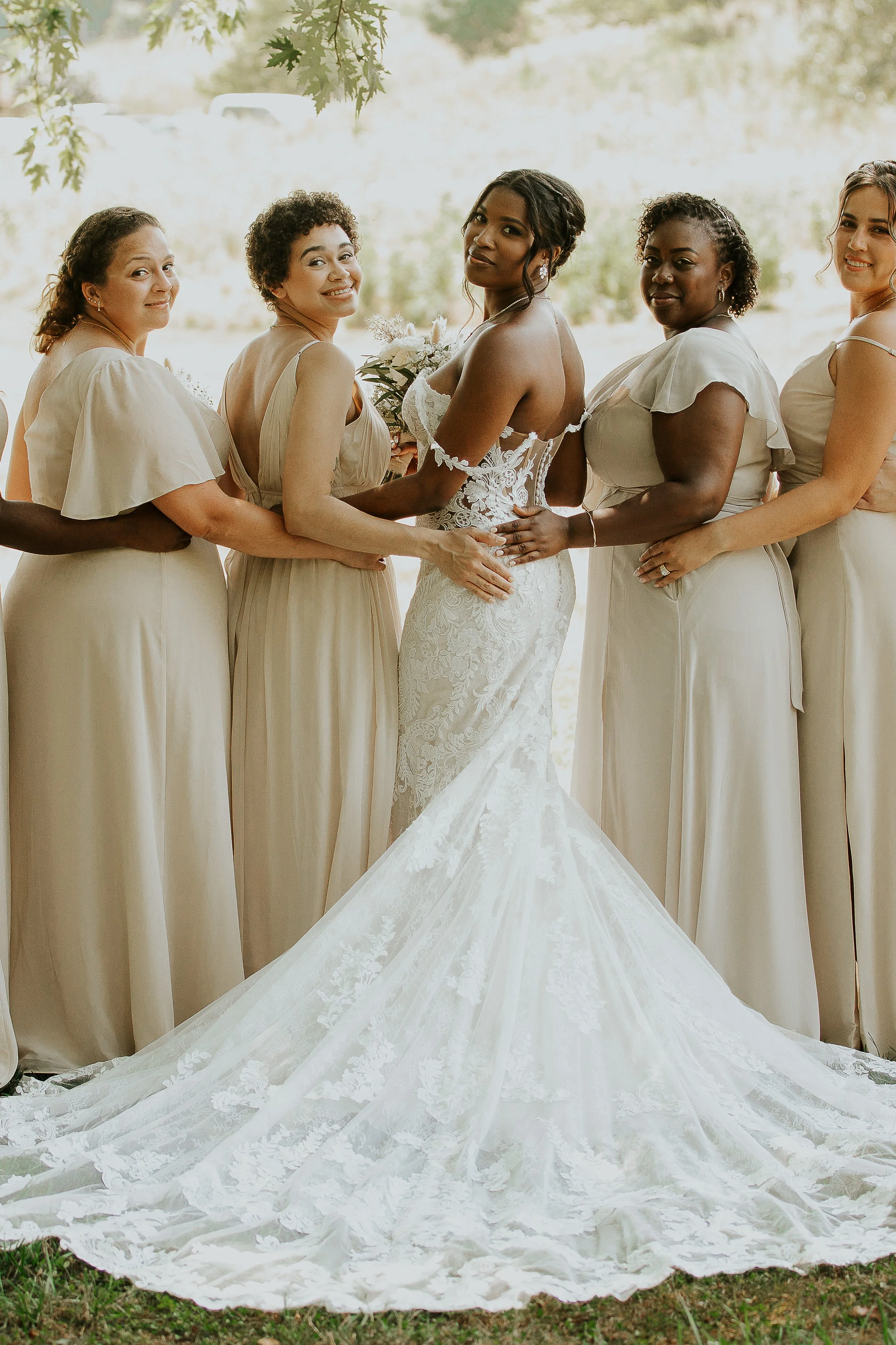 A group of women dressed in beige and white dresses, standing outdoors under trees, with the bride at the center wearing a white lace wedding gown with a train.