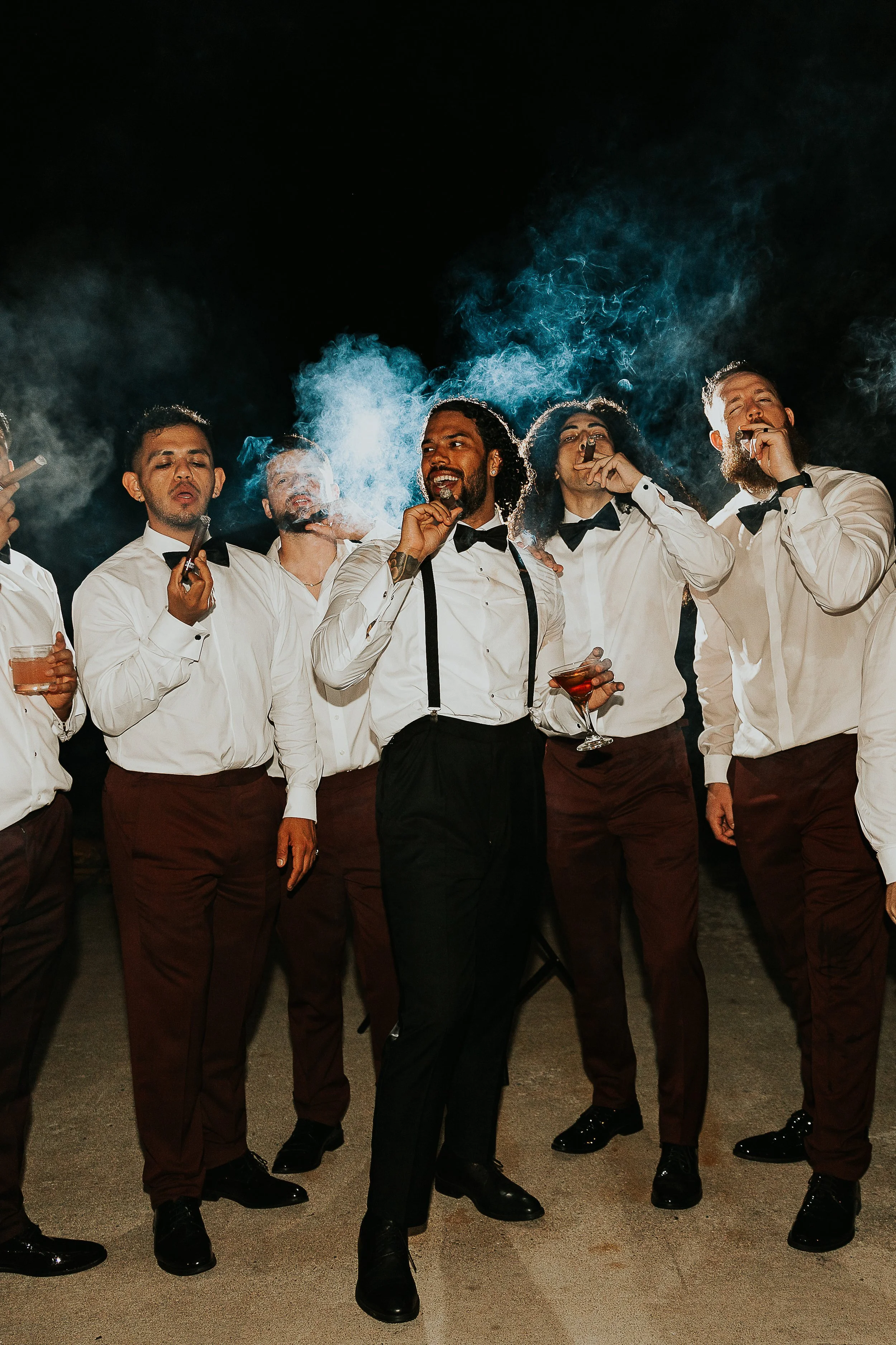 Group of men dressed in white shirts, black bow ties, and brown pants, holding cigars and drinks, standing together at night with some smoke in the air.