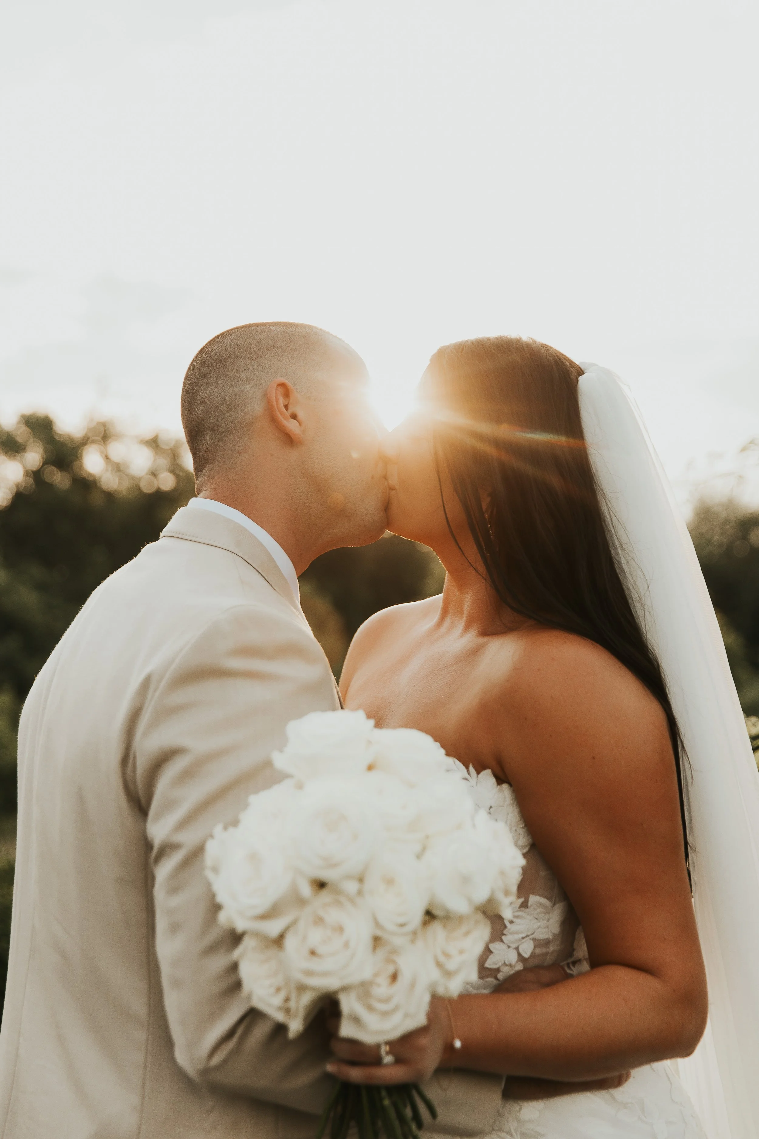A bride and groom kissing outdoors during sunset, with the bride holding a bouquet of white roses.