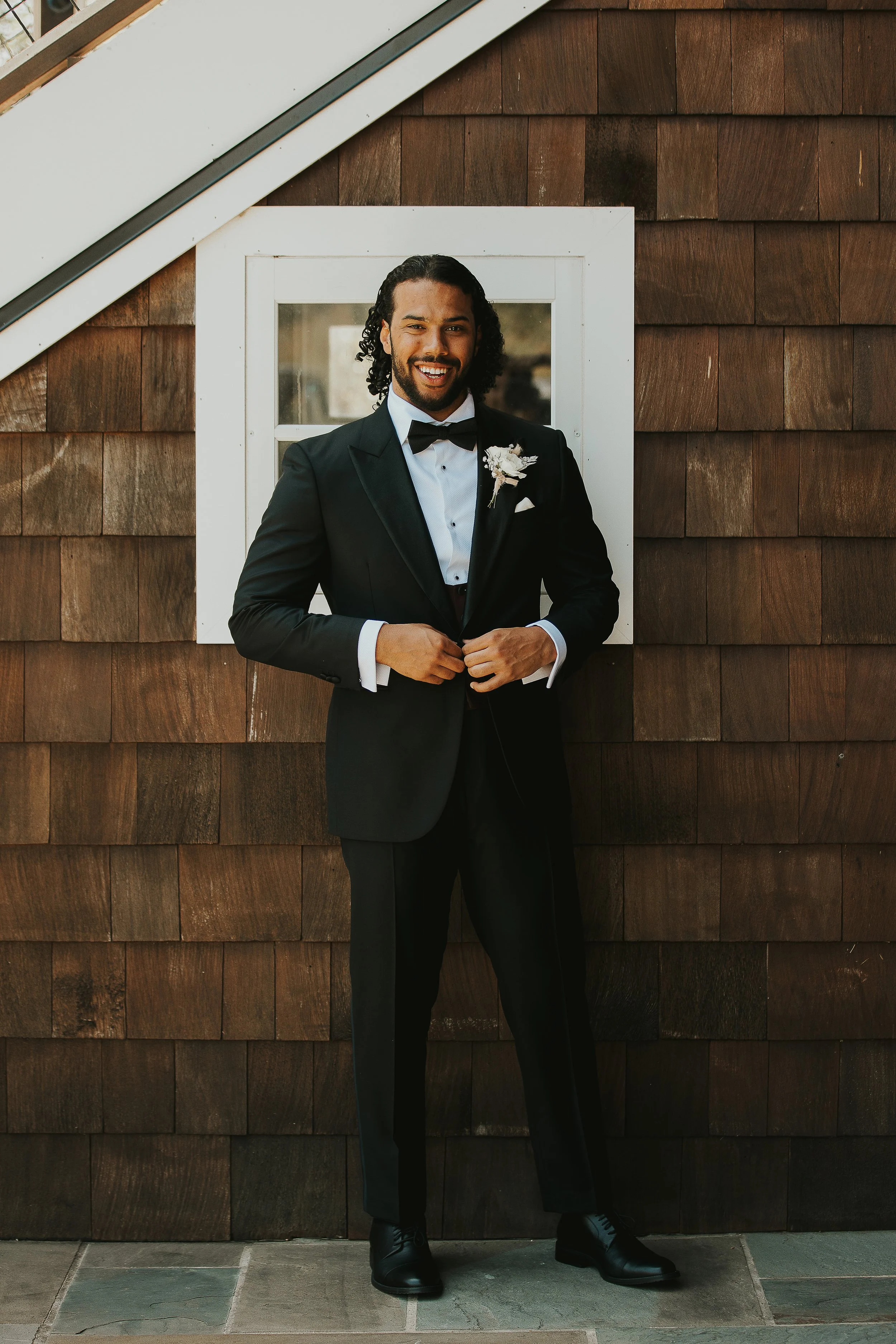 A man in a tuxedo with a bow tie and boutonniere standing in front of a wooden wall with a window, smiling at the camera.